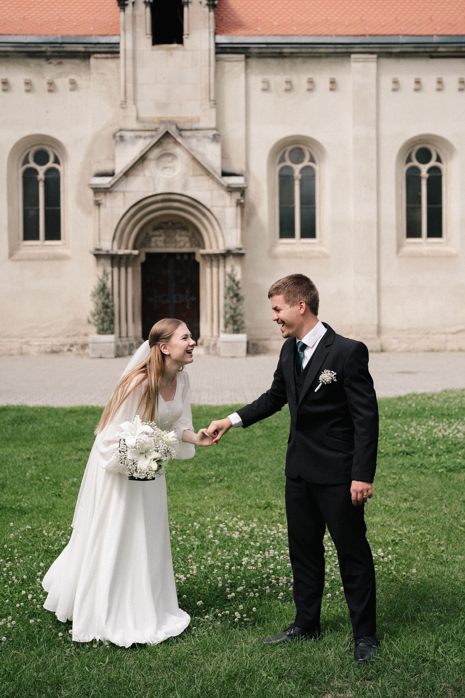 Playful wedding couple poses on grass in front of historic church building.