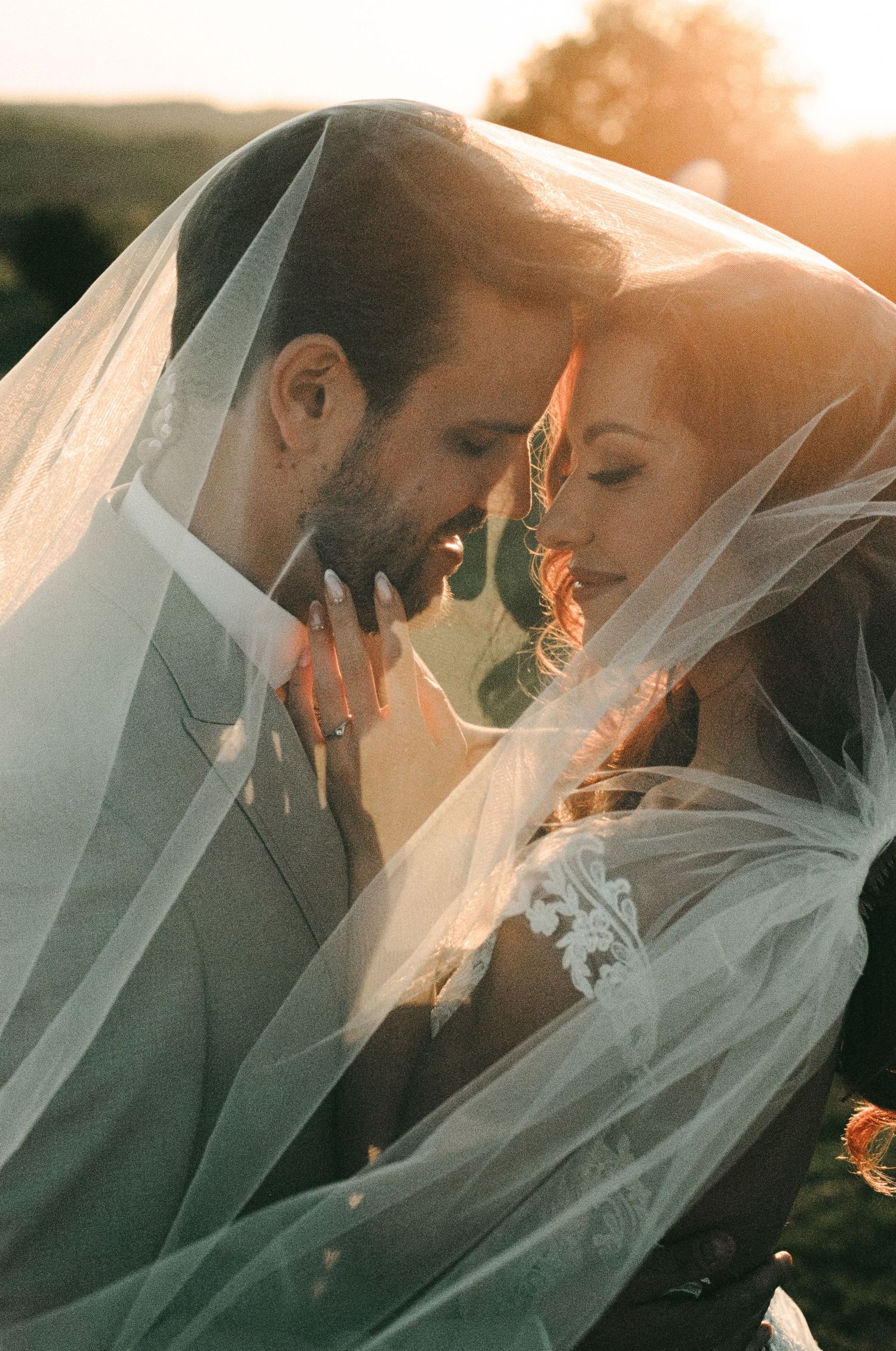 Bride and groom share an intimate moment under a flowing veil, bathed in golden sunset light.
