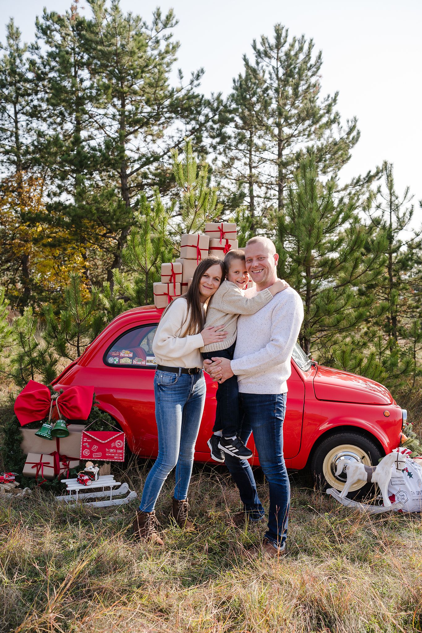 Family of three posing by a red vintage car loaded with Christmas gifts in a pine forest.