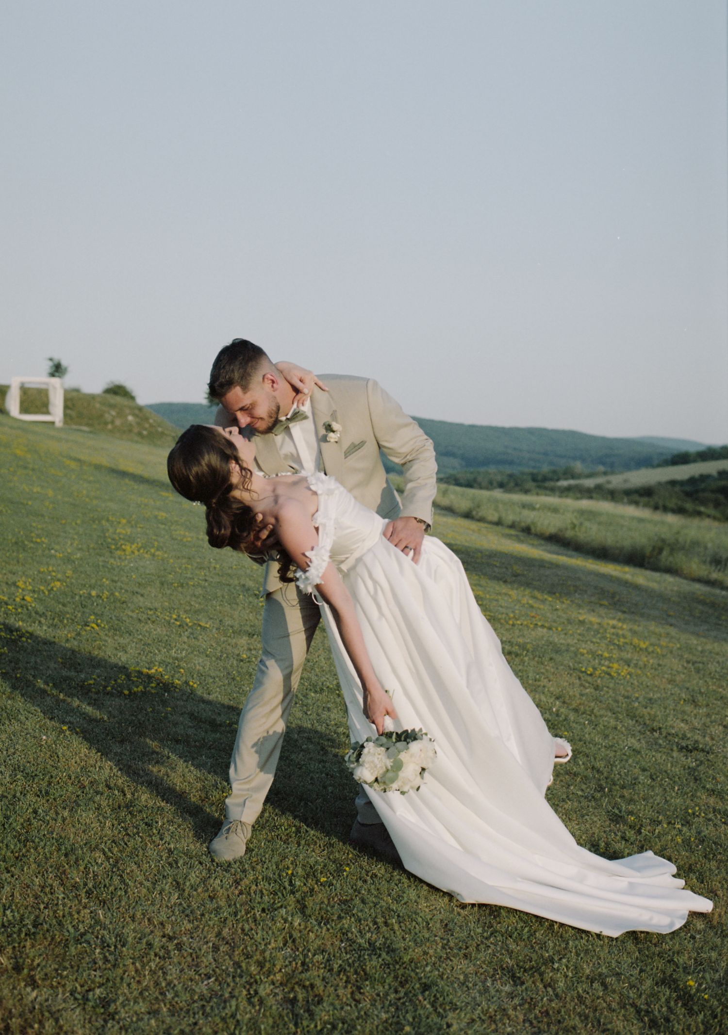 Romantic dip pose on a grassy hillside during golden hour wedding photoshoot.