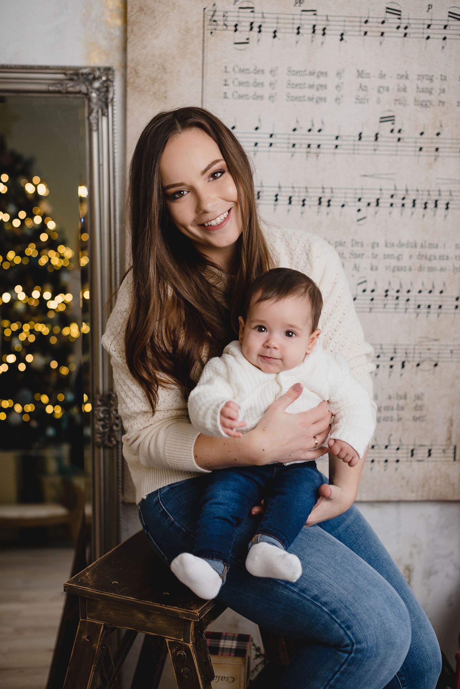 Smiling young mother holding baby in white sweater, Christmas tree bokeh lights and sheet music wall backdrop.