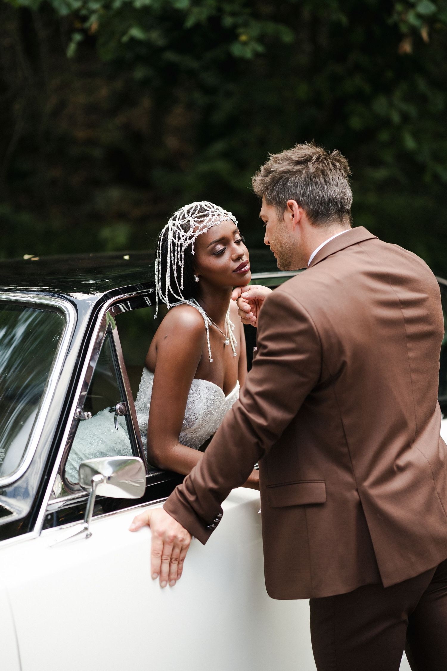 Interracial wedding couple sharing a romantic moment by a vintage white car, bride in lace gown with pearl headpiece.