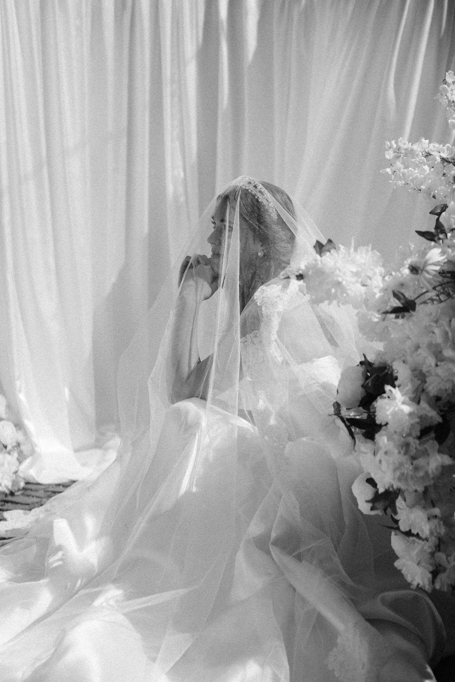 Black and white photo of a bride in a flowing veil and gown seated near white floral arrangements and draped curtains.