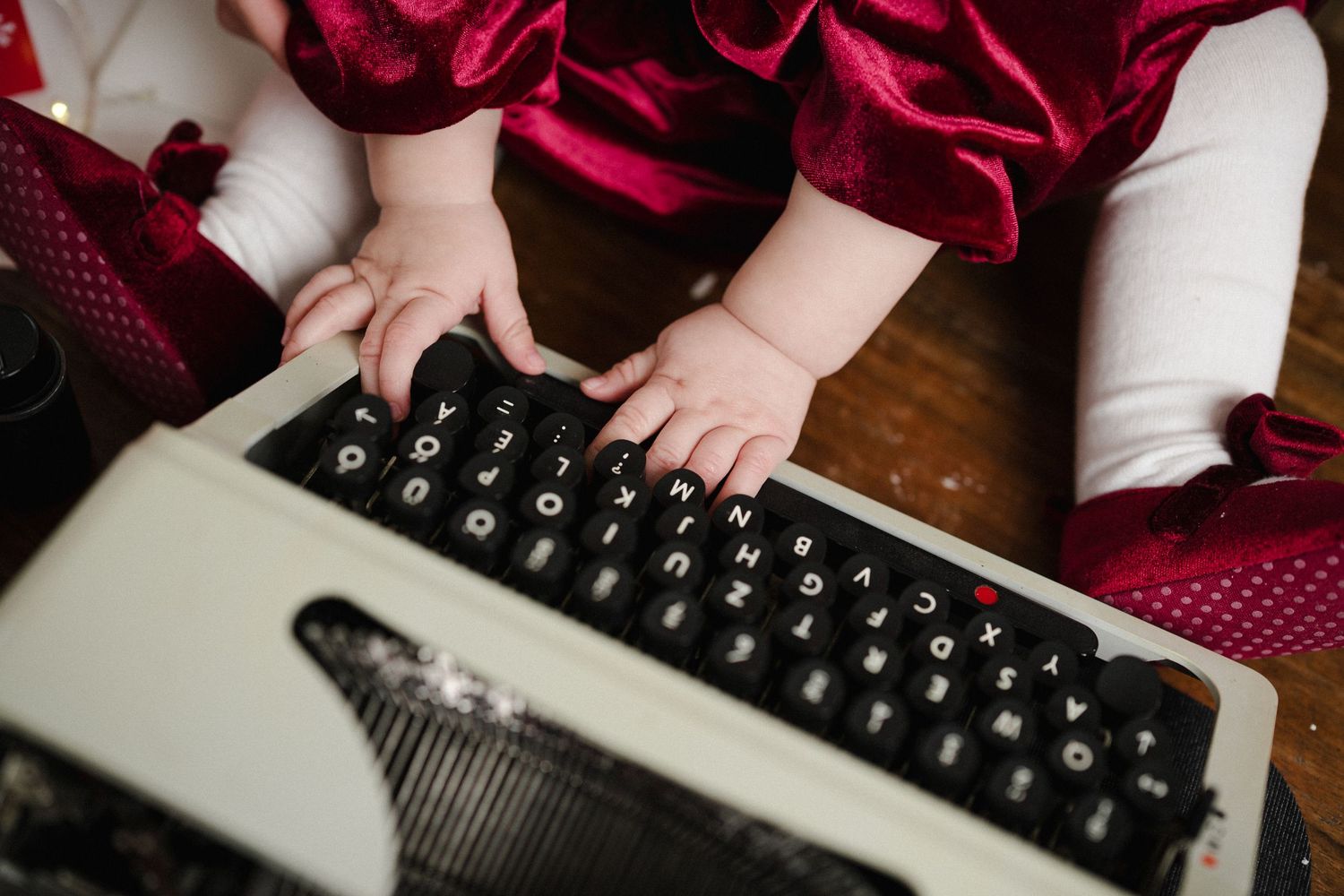 Baby hands typing on a vintage black and white typewriter keyboard, wearing a red velvet outfit.
