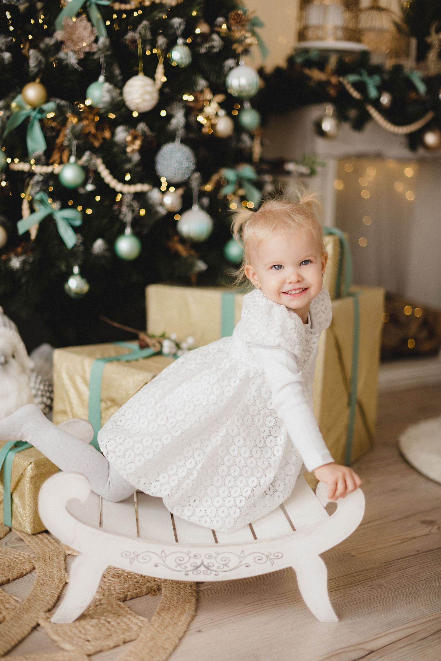 Smiling toddler girl in white dress sitting on wooden rocking chair near decorated Christmas tree with gold gifts.