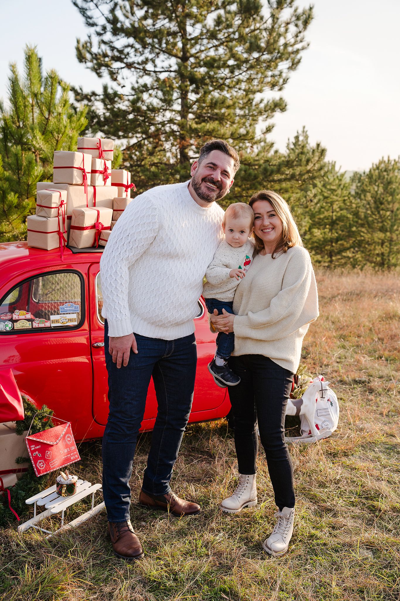 Happy family with baby posing by a red vintage car loaded with Christmas gifts in a pine forest setting.
