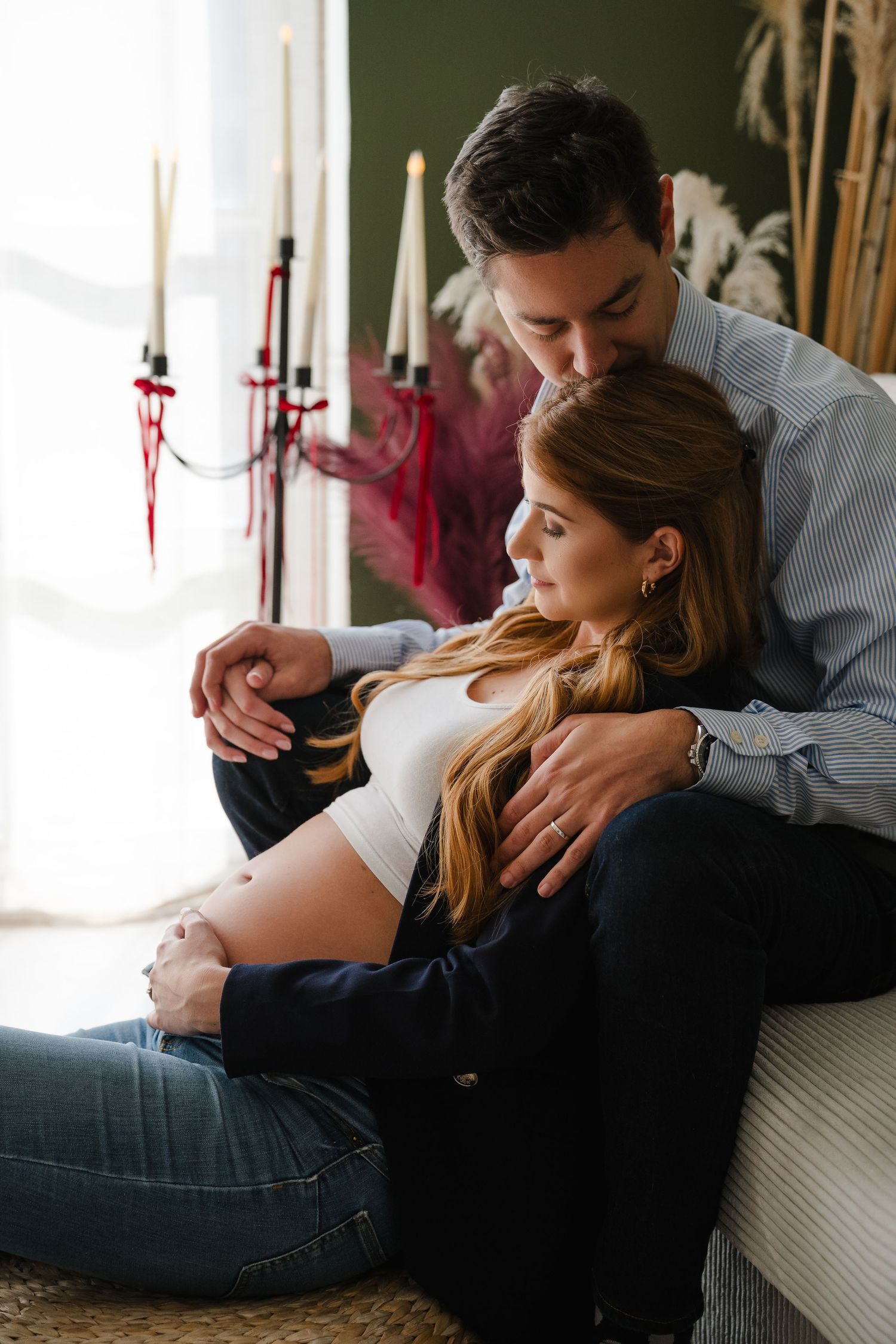 A couple sits together on the floor, the man embracing his pregnant partner while gently kissing her head near candlelight.