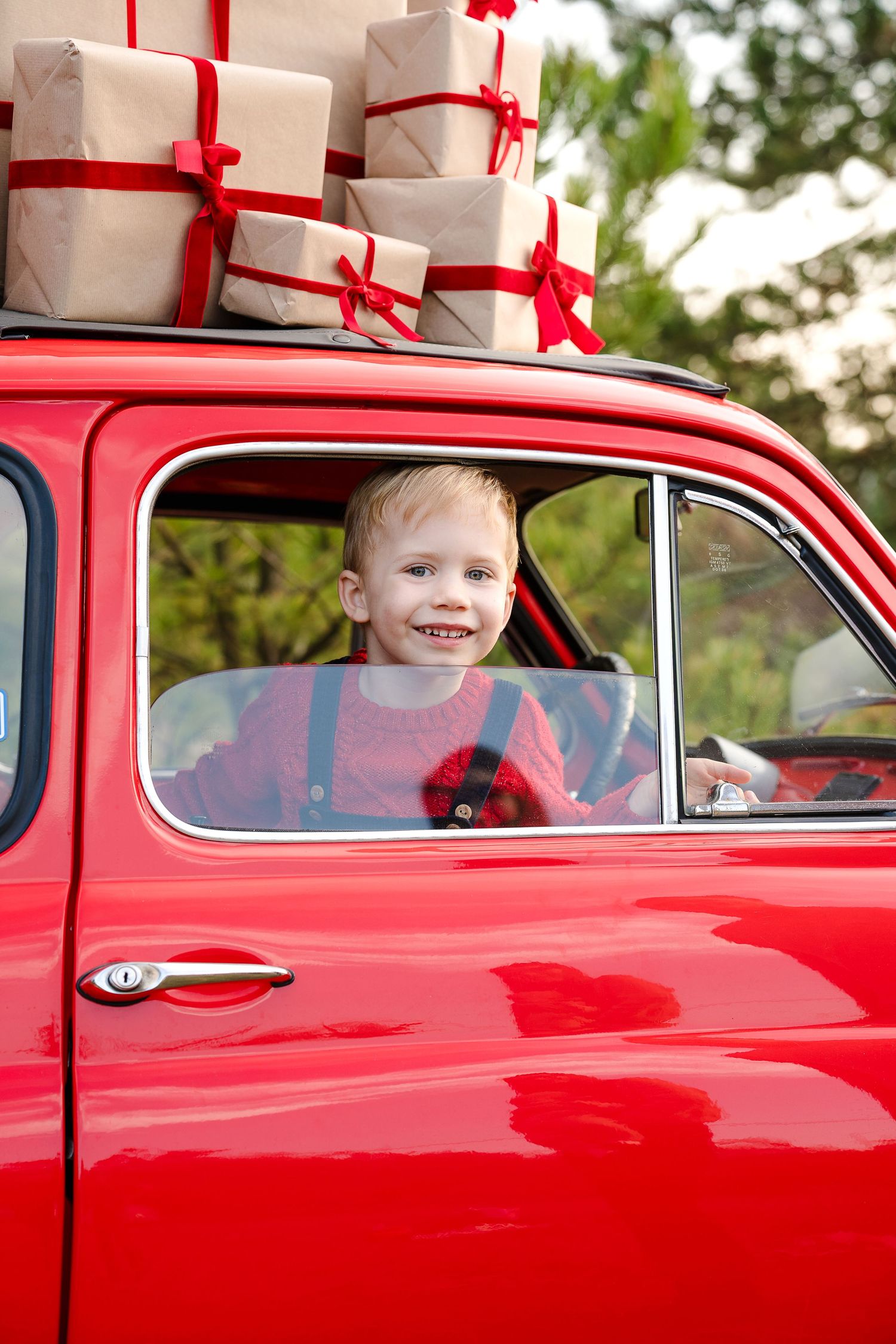 Smiling blonde boy sitting in a vintage red car with gift boxes tied with red ribbons stacked on the roof.