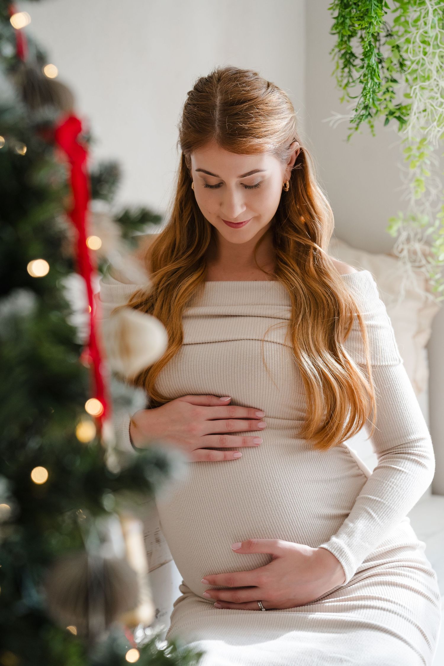 Pregnant woman in white dress tenderly holding her baby bump near a decorated Christmas tree with red ribbons.