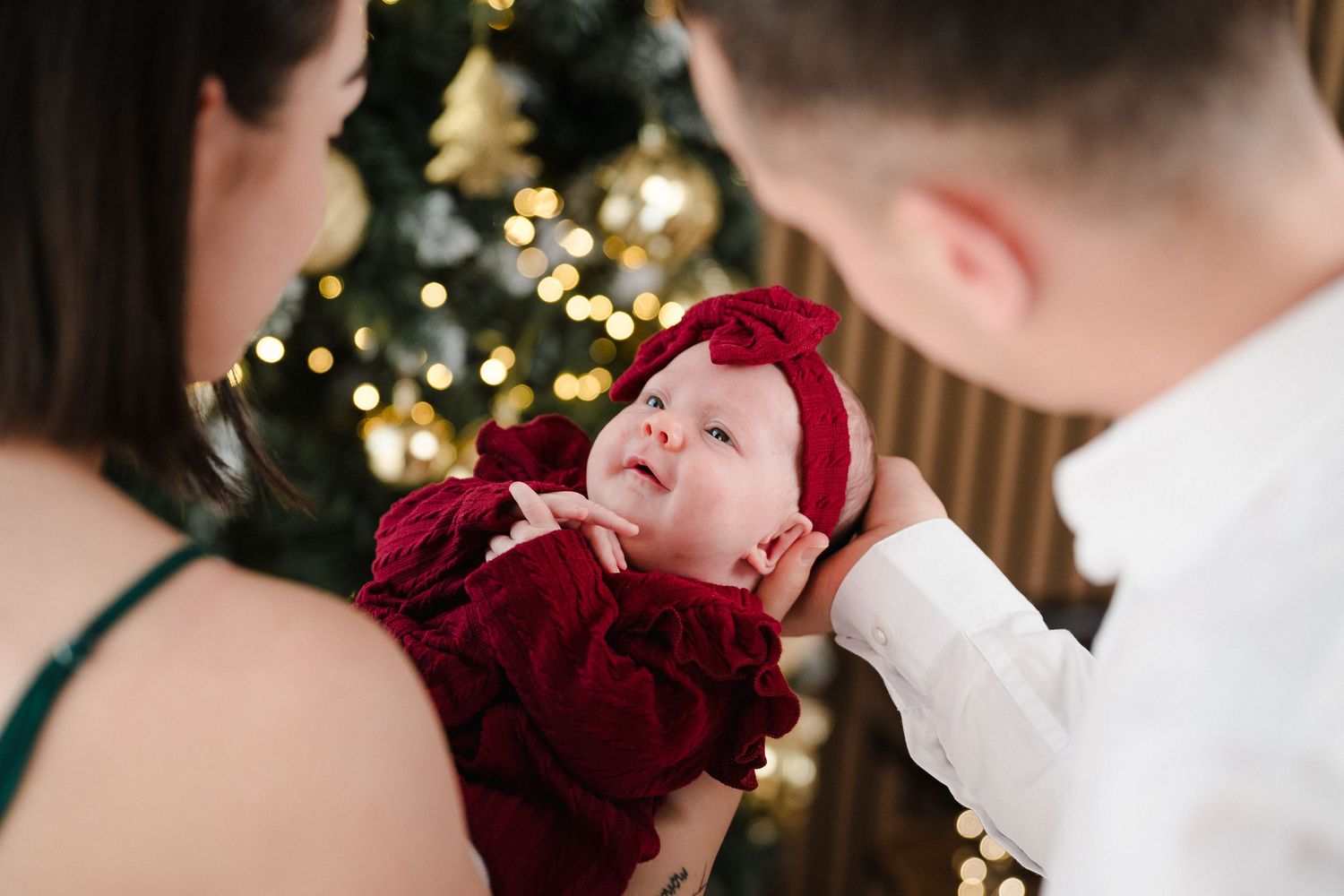 Parents lovingly gazing at their baby dressed in red velvet outfit near a decorated Christmas tree.