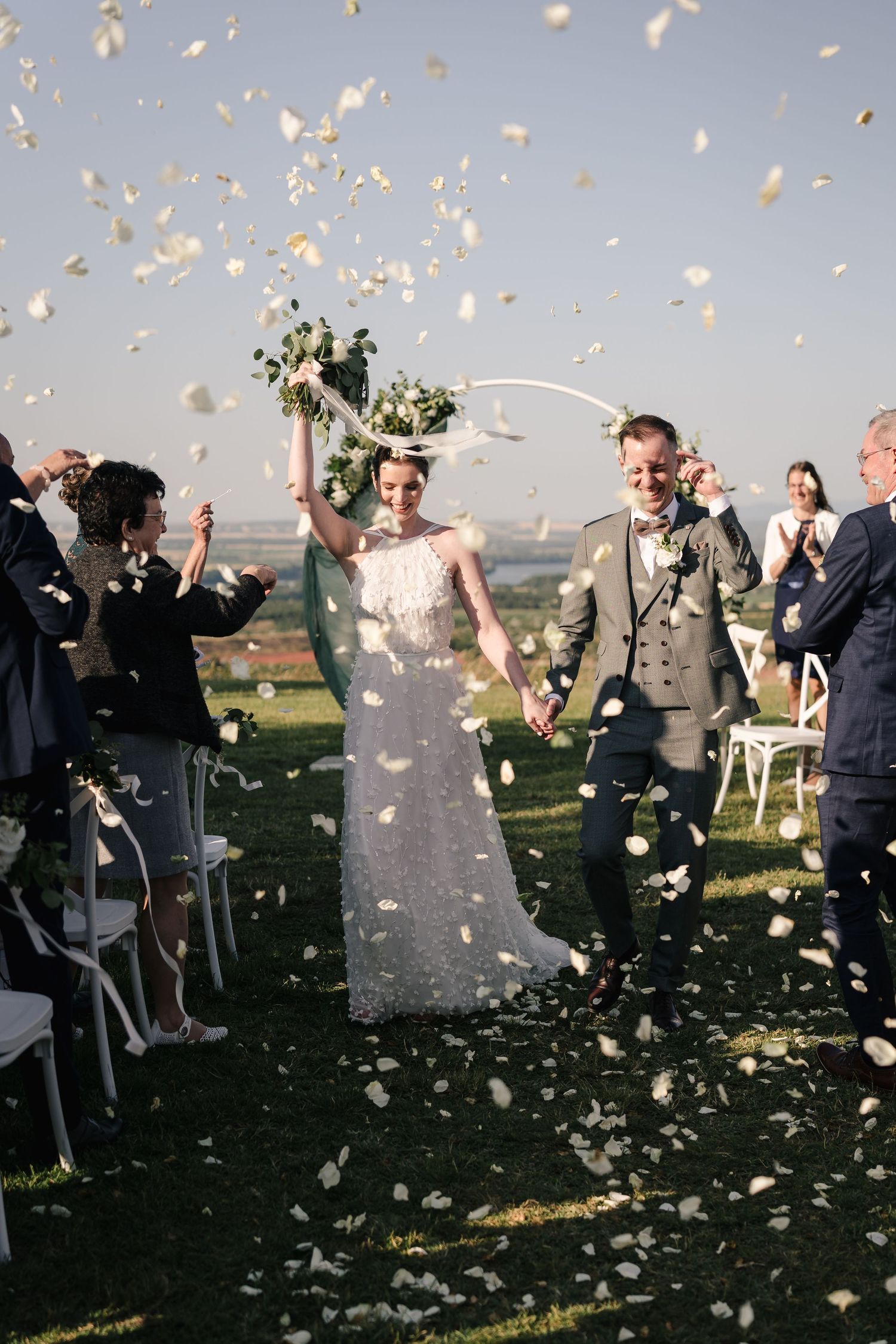 Wedding guests celebrate by tossing white flower petals into the air during an outdoor ceremony.
