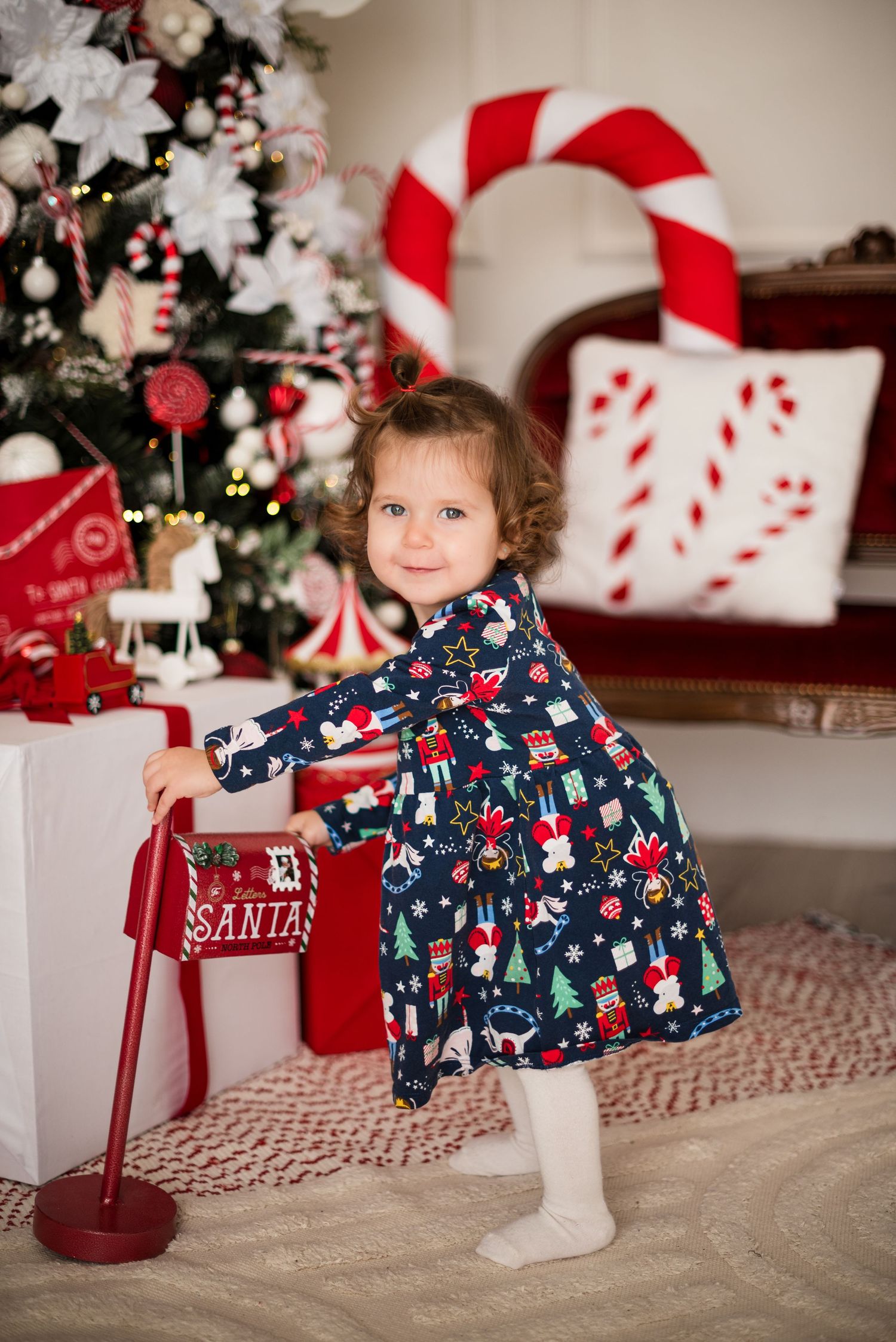 Toddler girl in Christmas dress mailing letter to Santa beside decorated tree with candy cane decorations.