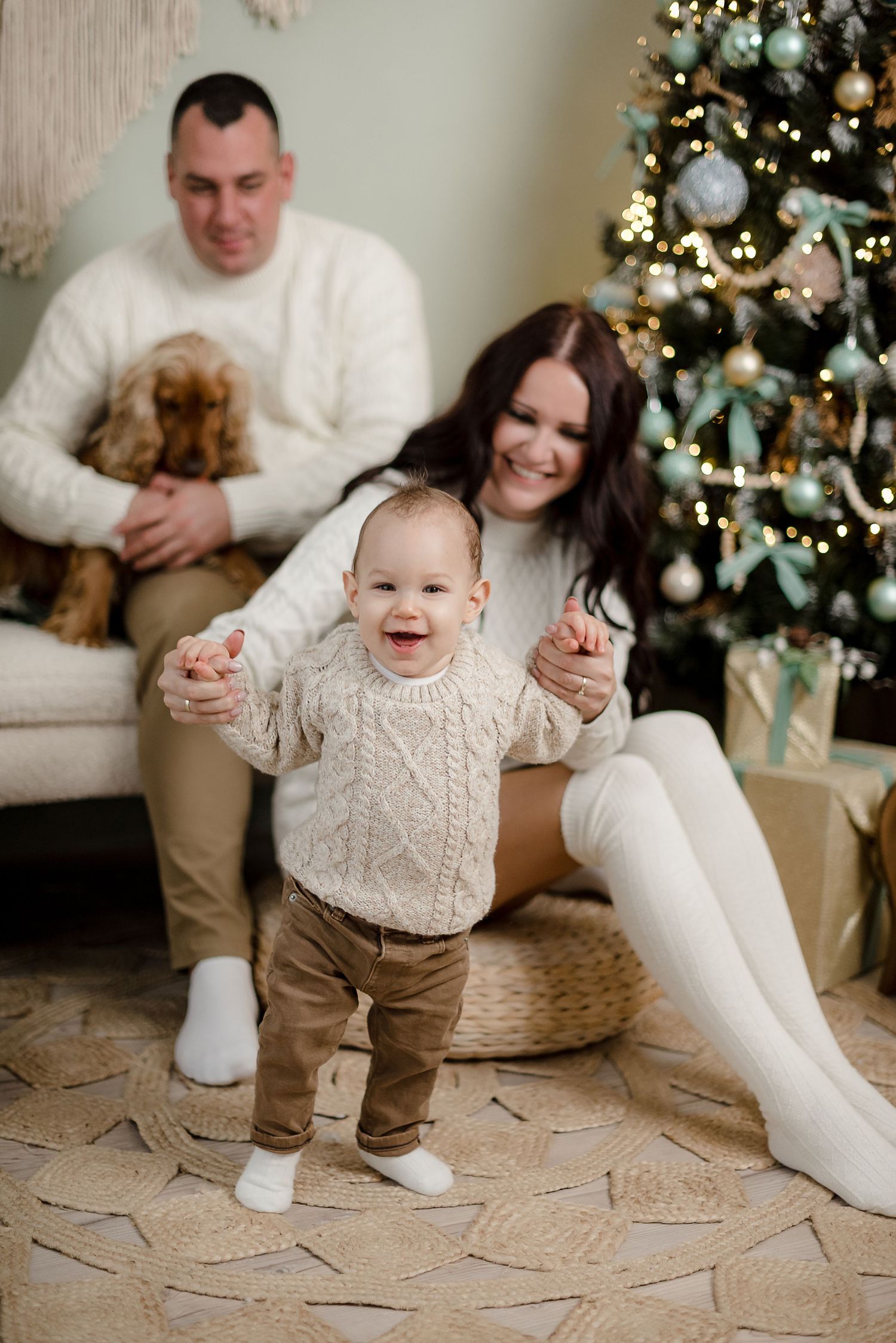 Happy family with baby taking first steps near Christmas tree, parents in white sweaters holding toddler hands with dog nearby.