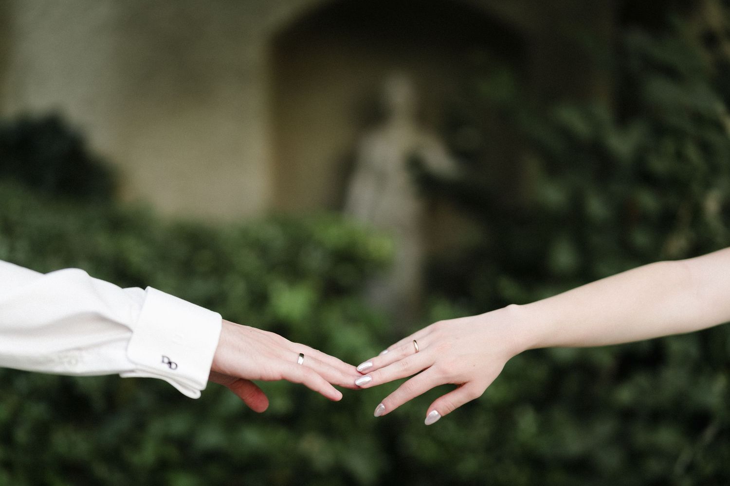 Two hands reaching toward each other outdoors, with a blurred statue and lush green ivy in the background.