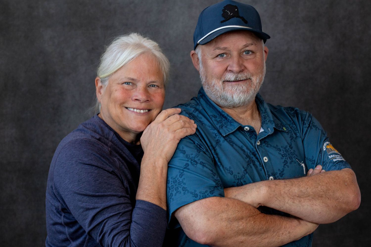 A smiling couple in blue clothing pose together for a professional portrait against a dark background.
