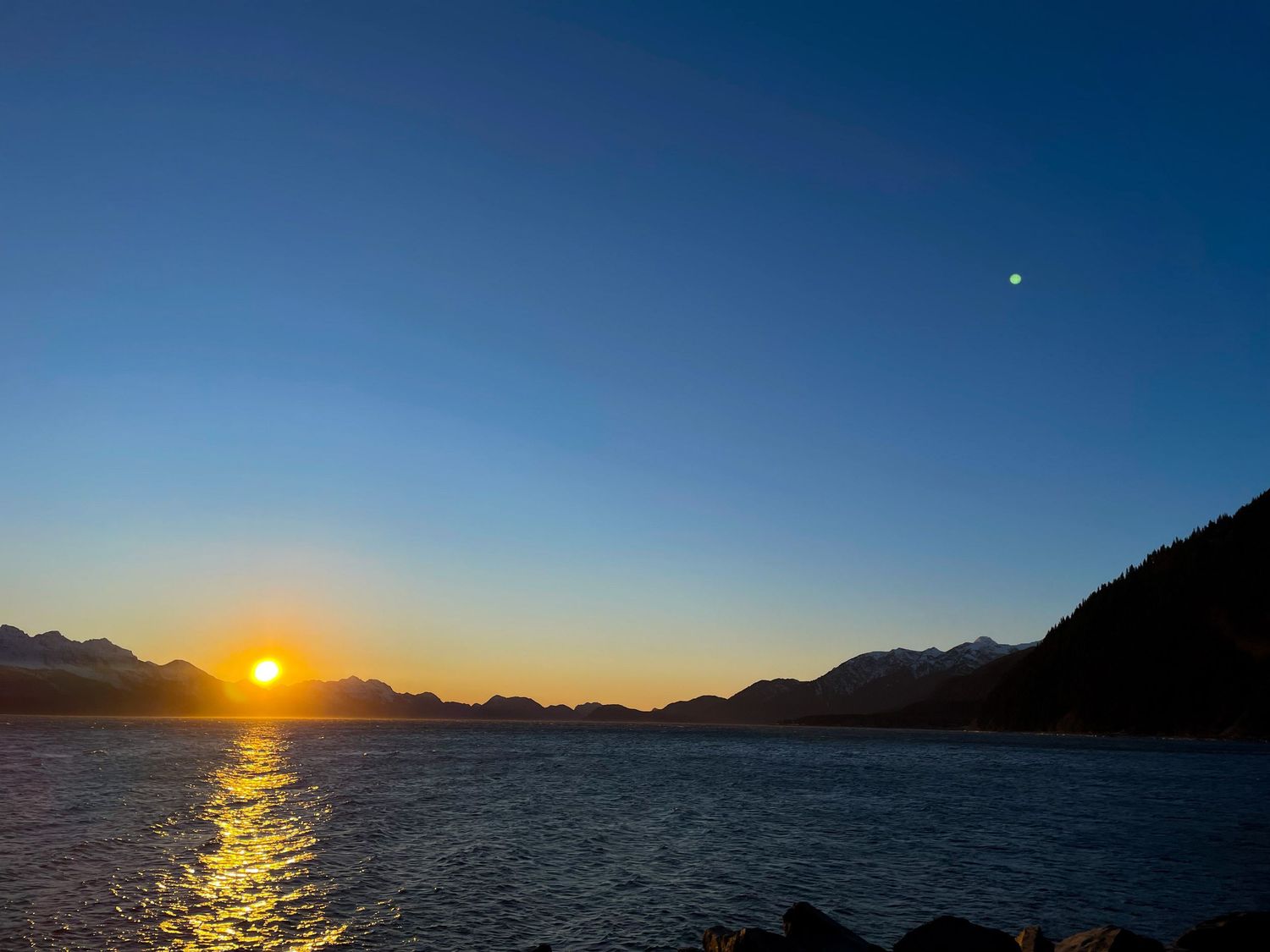 A stunning sunset over calm ocean waters creates a golden reflection path against a deep blue sky and mountain silhouettes.