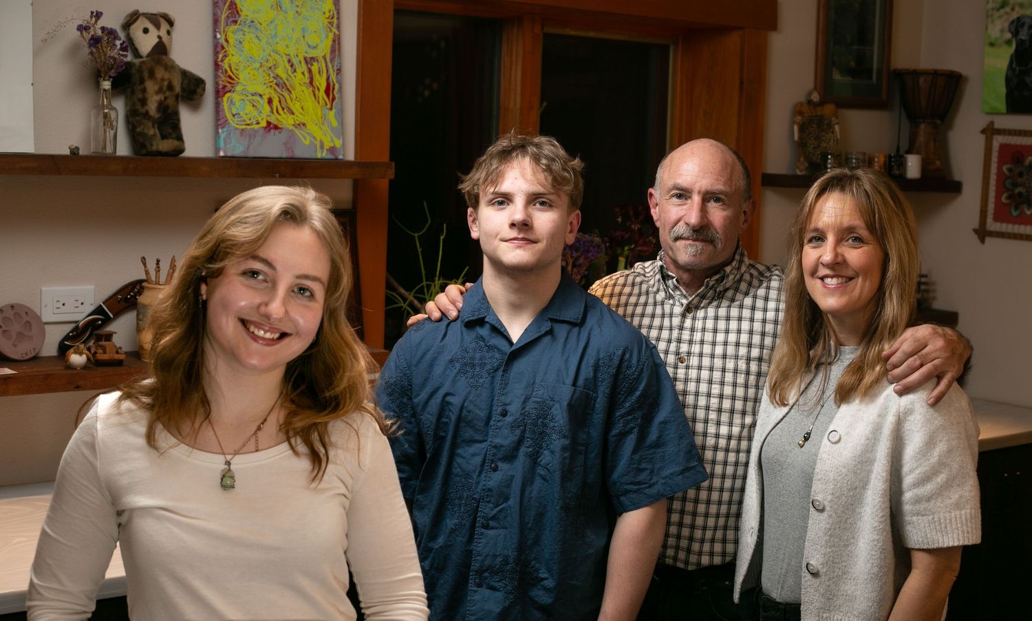 A family of four poses together in their home with wooden shelves and decorations visible in the background.