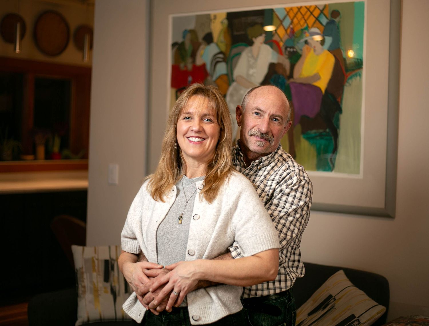 Two chefs embrace and laugh together in a restaurant setting with framed artwork on the wall behind them.