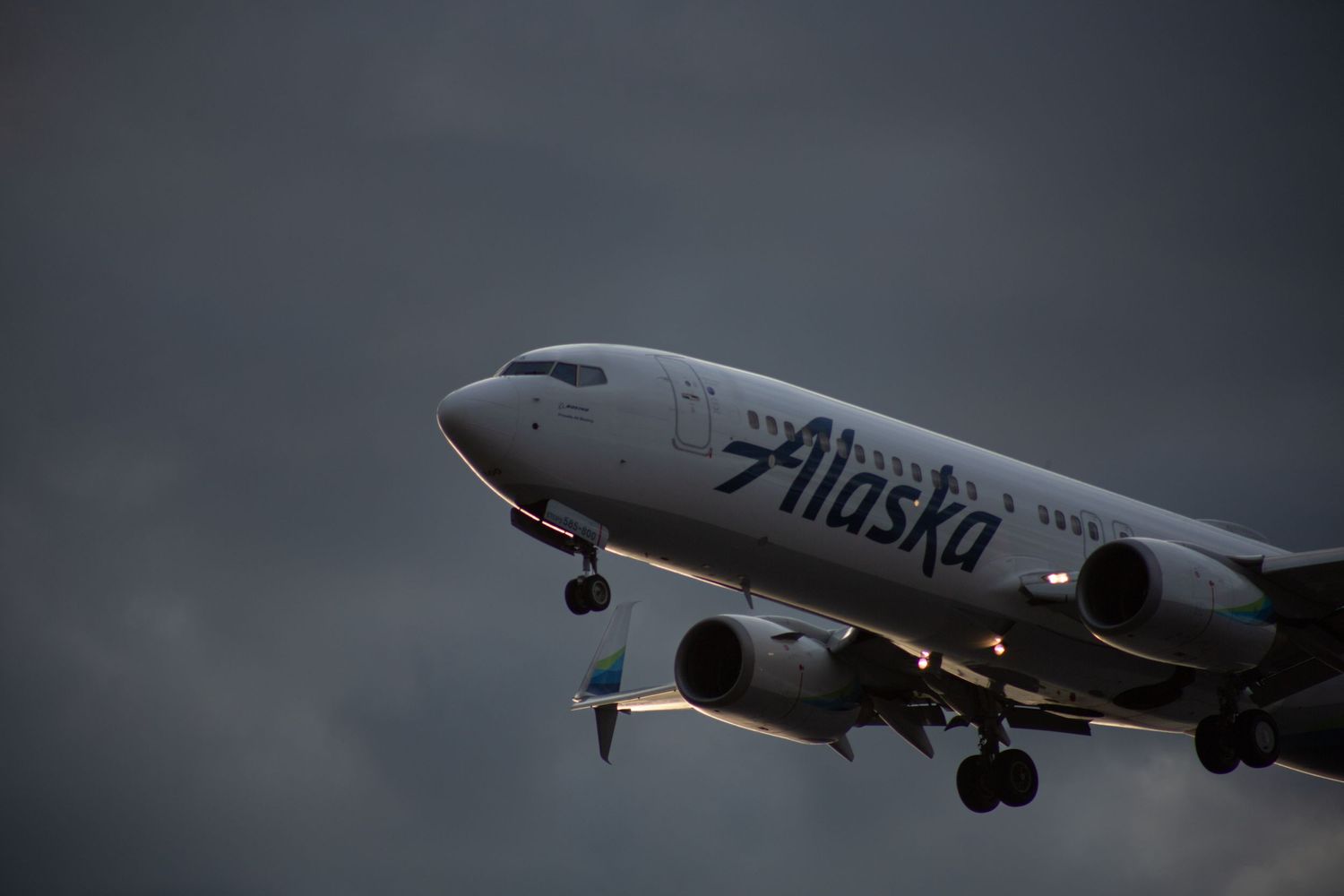 Alaska Airlines Boeing 737 aircraft on approach to land against a dramatic dark cloudy sky at dusk.