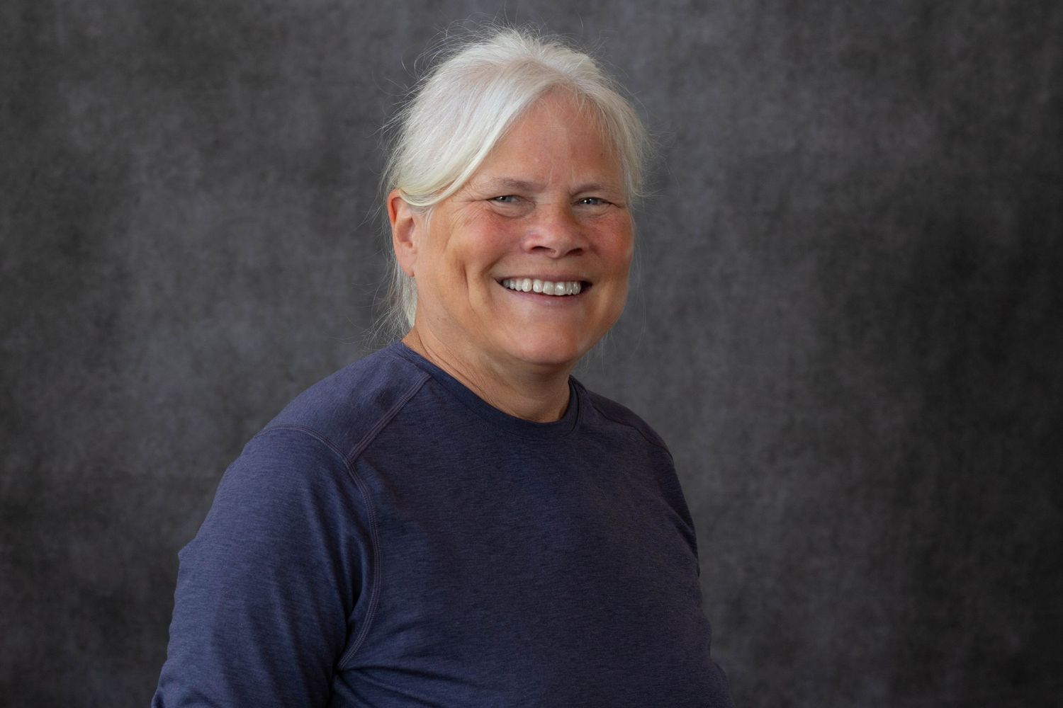 Professional headshot portrait of someone wearing a navy sweater against a dark gray background, smiling warmly.