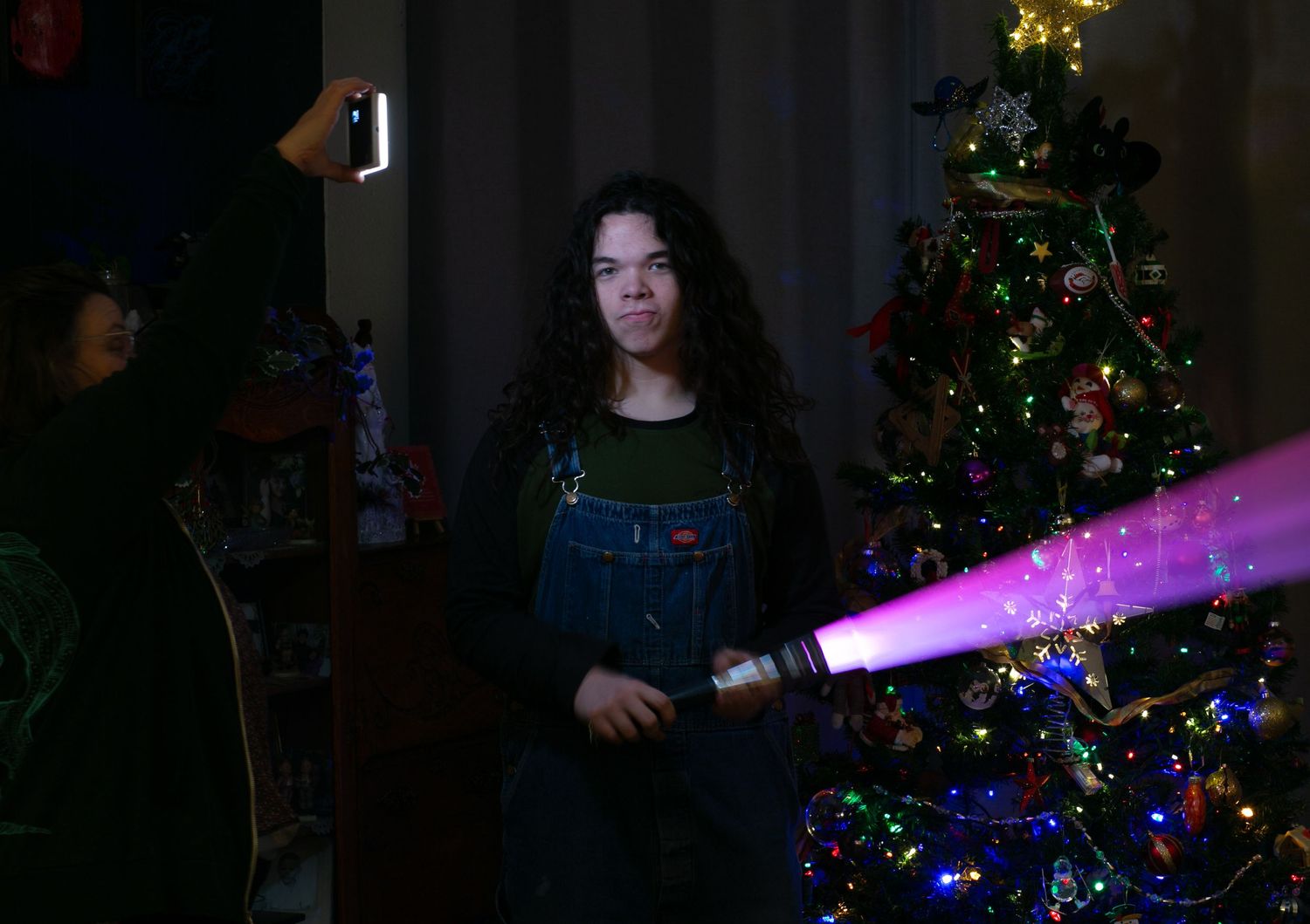 Someone in overalls stands next to a decorated Christmas tree with colorful lights in a dark room.