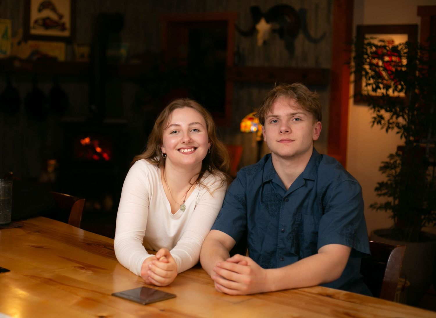 Two people sit together at a wooden table in a dimly lit room with warm lighting creating an intimate atmosphere.