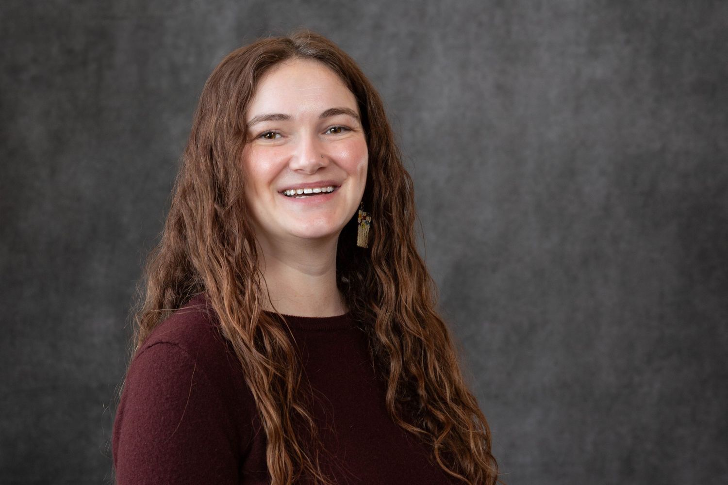 Professional headshot portrait with long wavy brown hair and burgundy sweater against dark gray background.