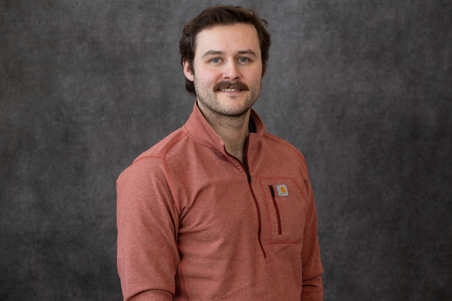 A person in a rust-colored Carhartt pullover smiles while posing against a dark gray textured background.