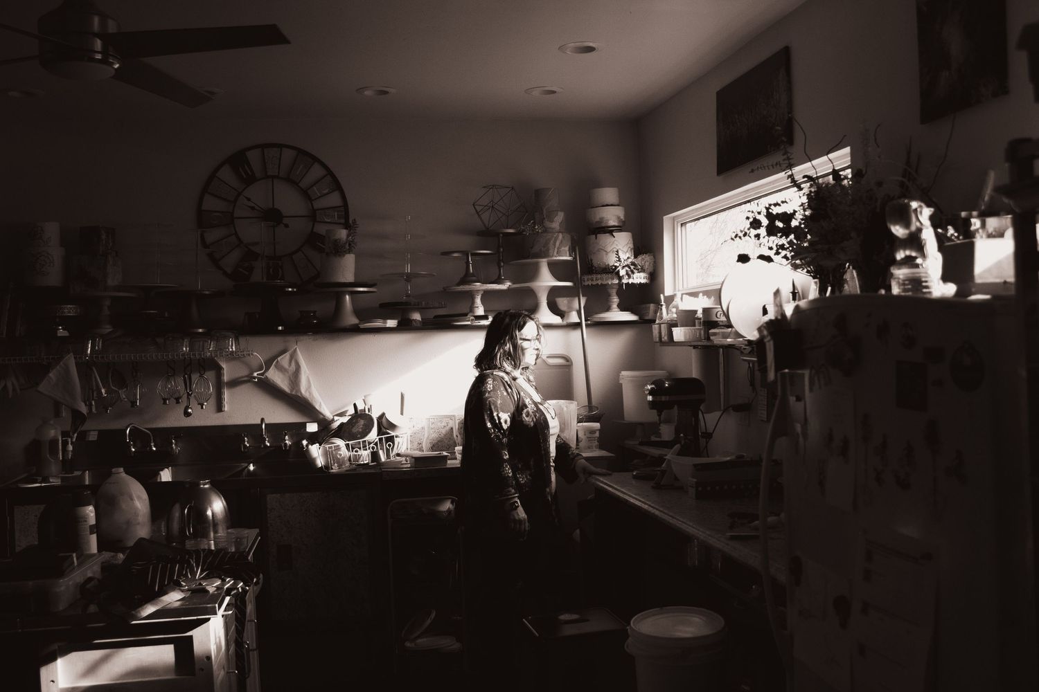 Black and white photo of a woman standing in a dimly lit vintage kitchen with a clock on the wall.