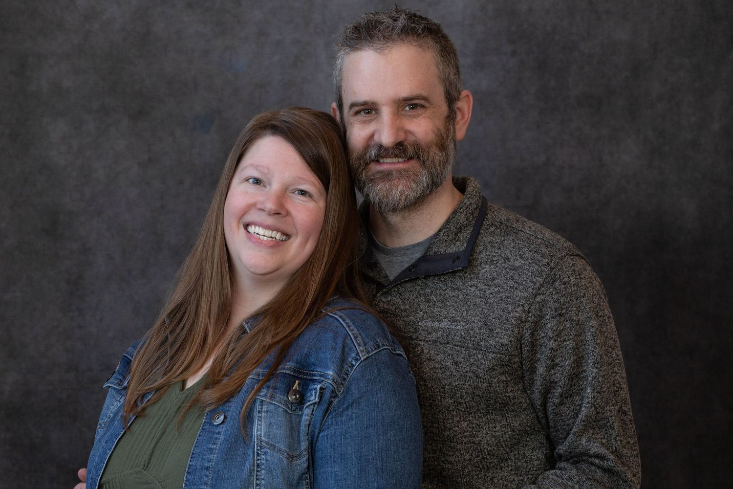 A couple poses together against a dark gray backdrop, wearing casual clothing and warm smiles.