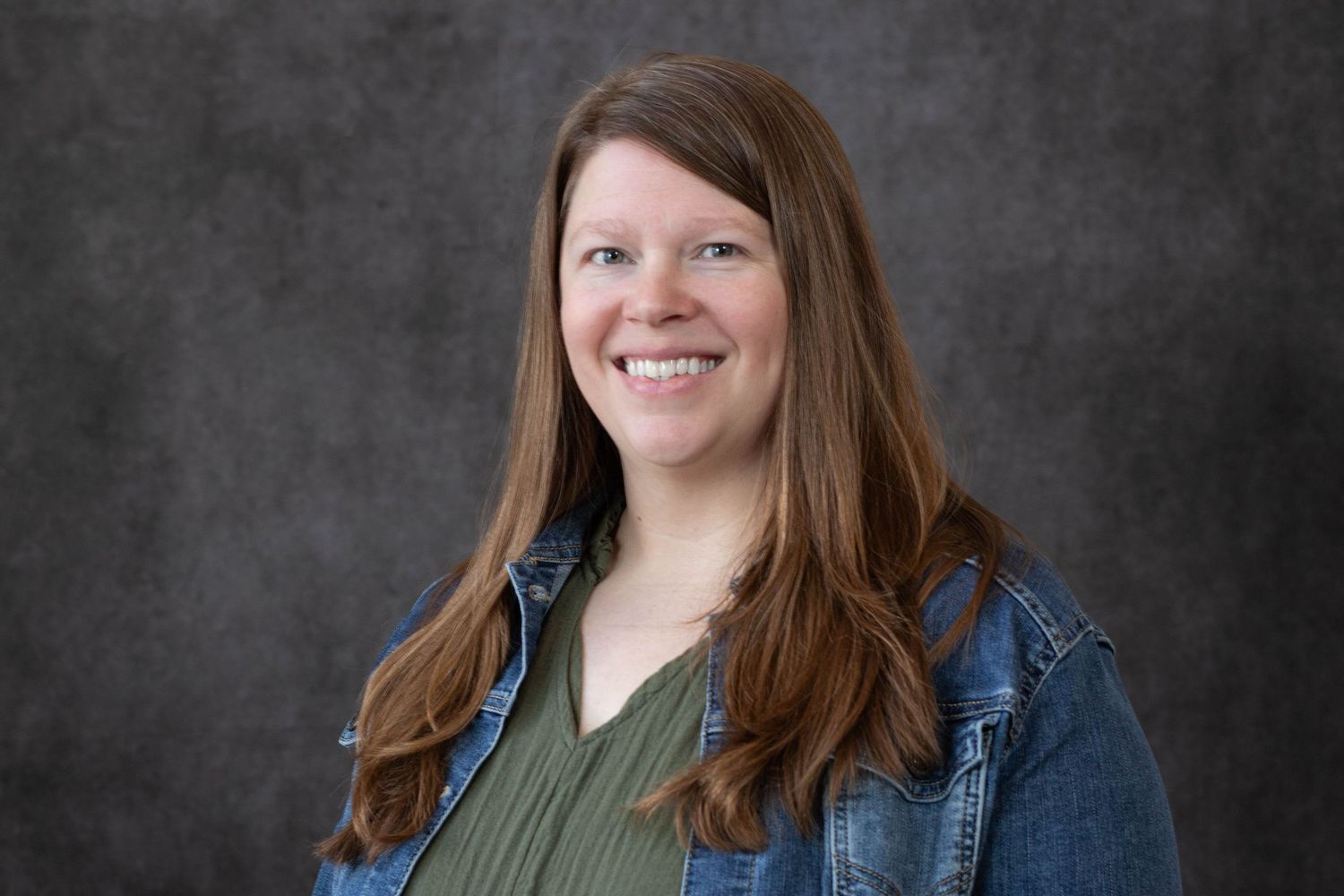 Professional headshot of someone wearing a denim jacket and olive green top against a dark gray background.