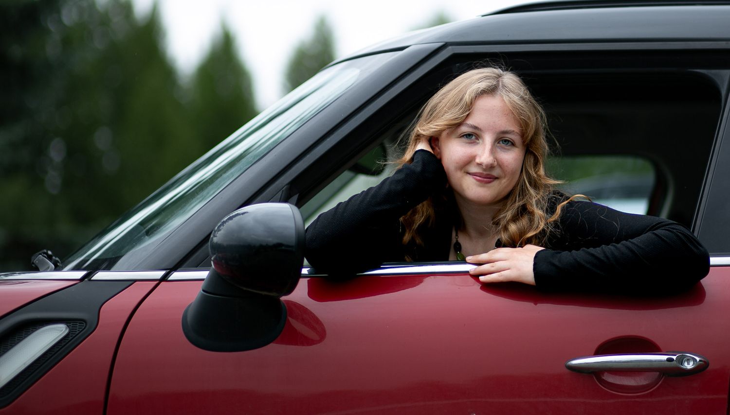 Someone leans out the window of a shiny red car with trees visible in the background.