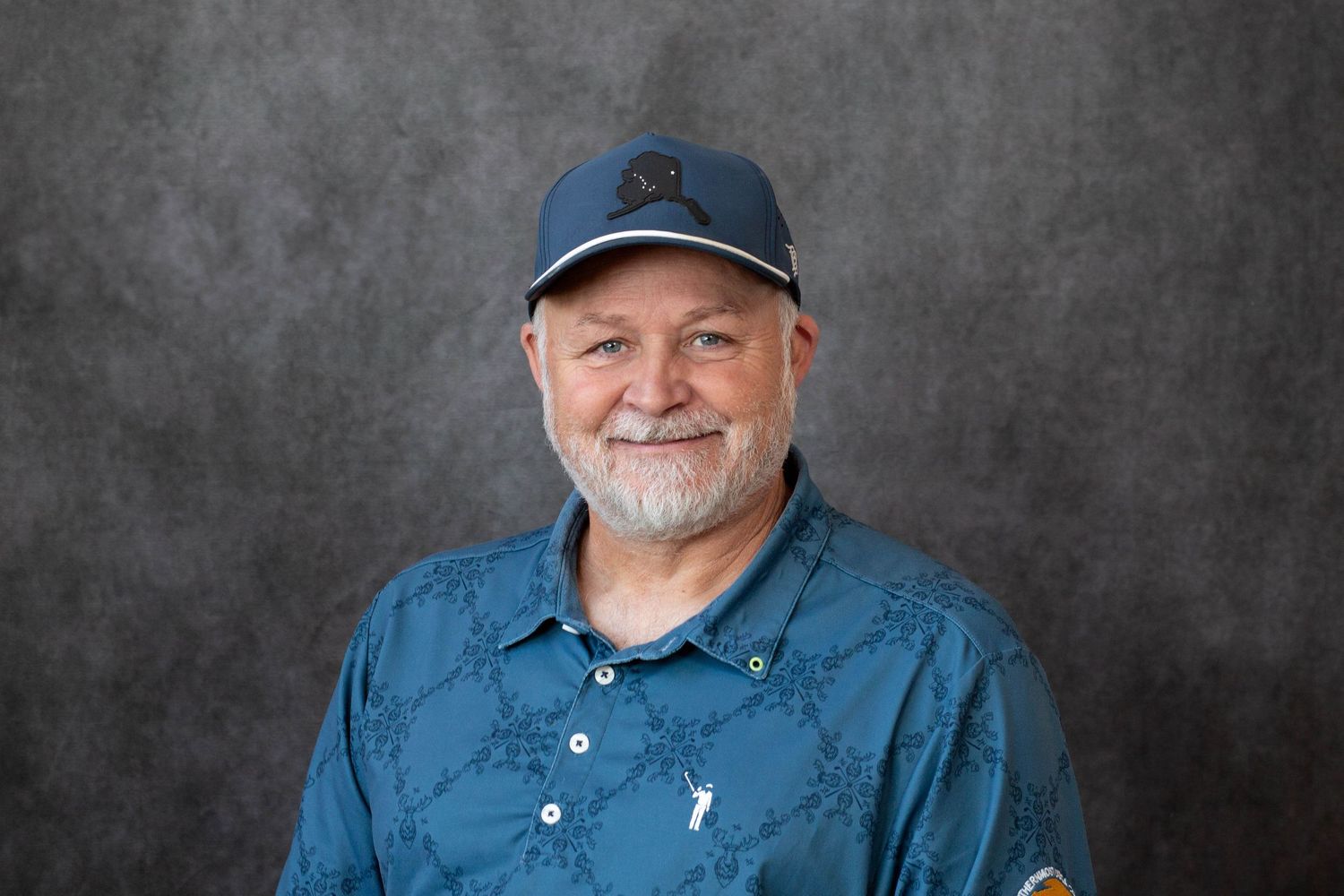 Professional studio portrait of someone wearing a blue baseball cap and denim shirt against a gray background.