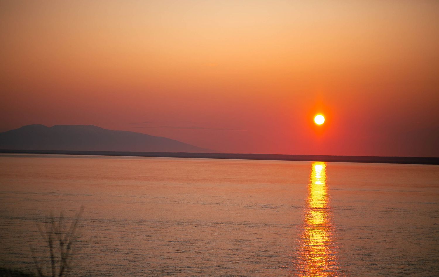 A vibrant orange sun sets over calm waters, casting a golden reflection path across the lake with mountains in the distance.