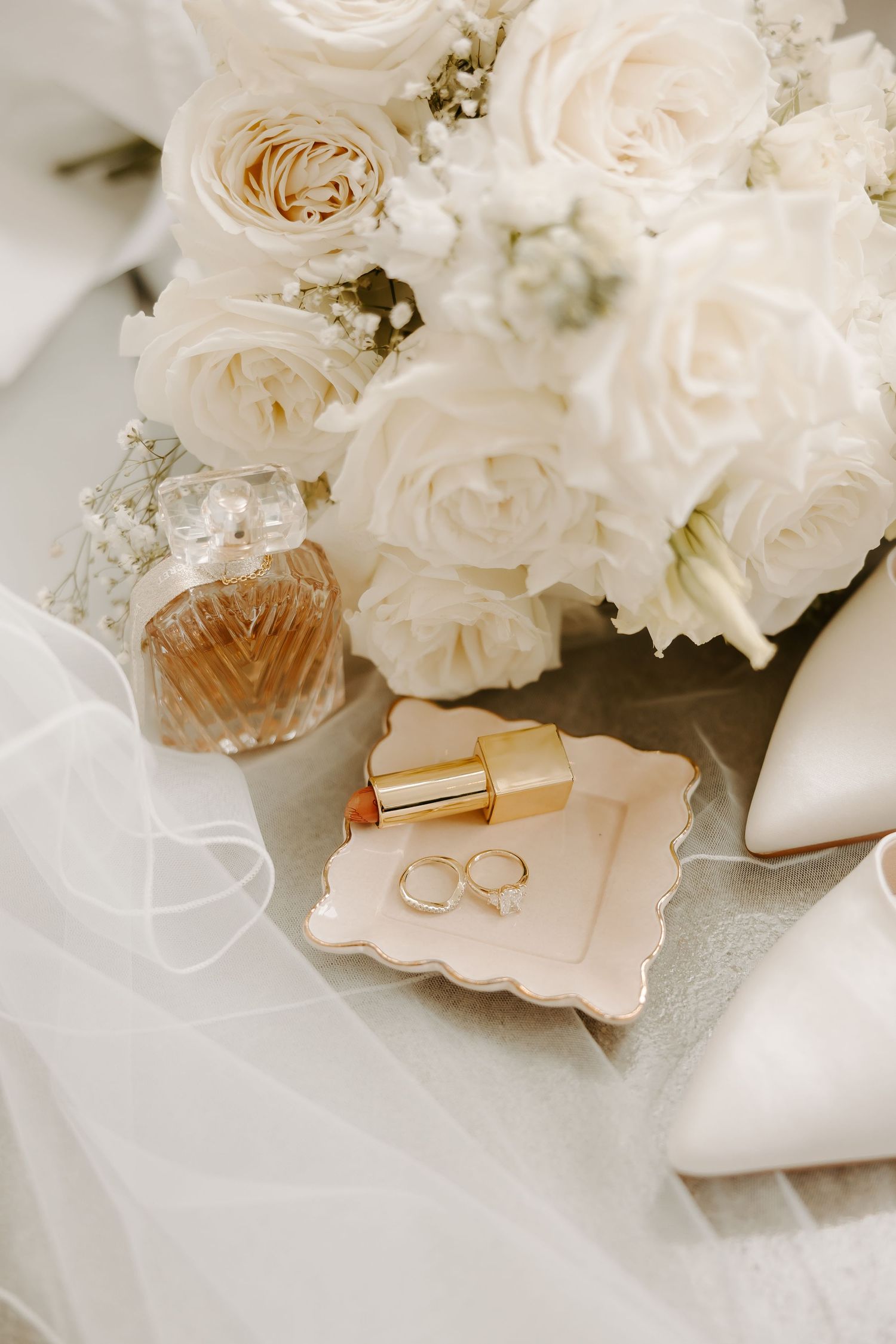 Elegant bridal flat lay with white roses bouquet, gold lipstick, wedding rings, perfume bottle, and white heels on soft fabric.