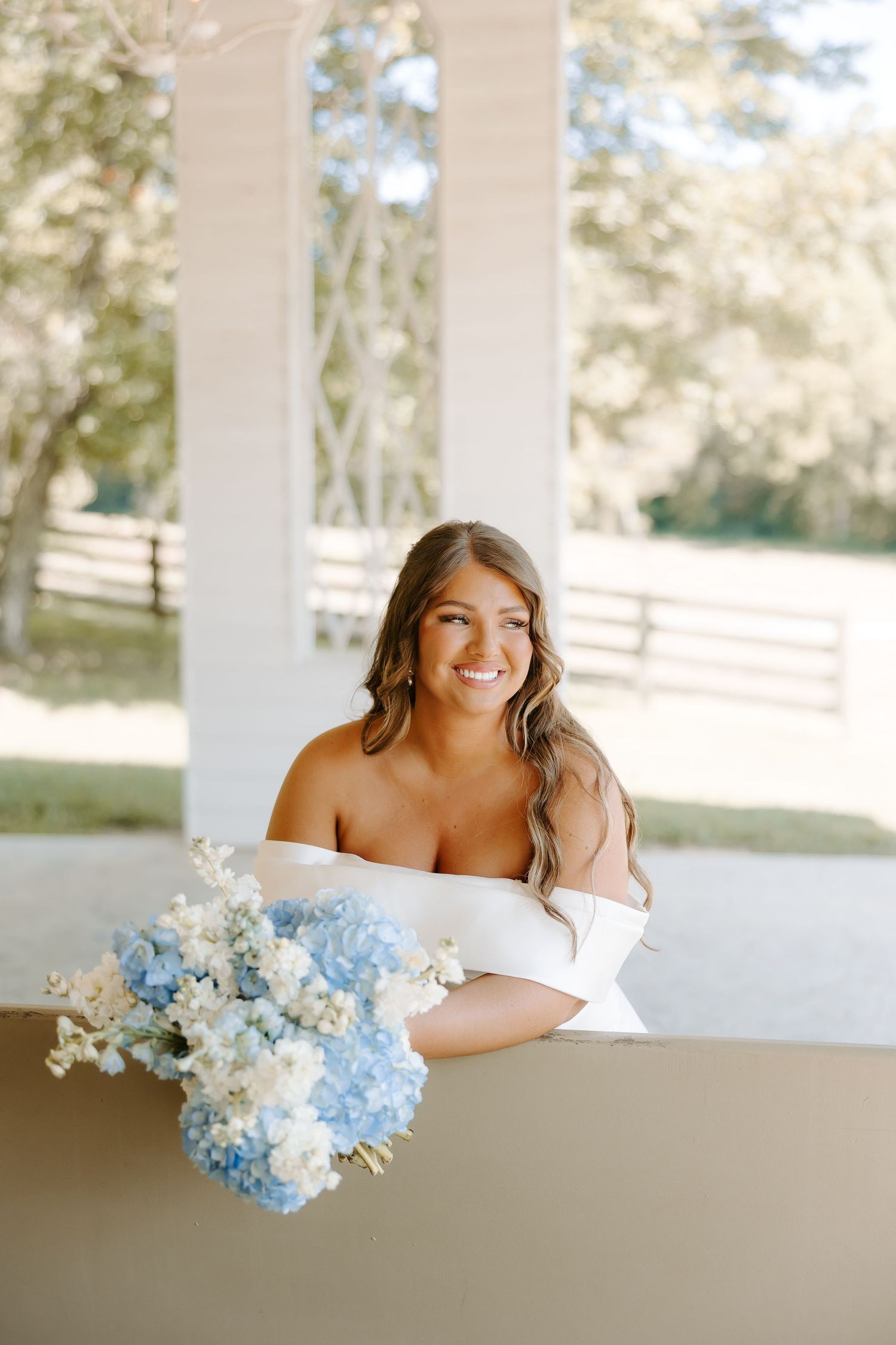 Smiling bride in off-shoulder white gown holding blue and white hydrangea bouquet at an outdoor venue.