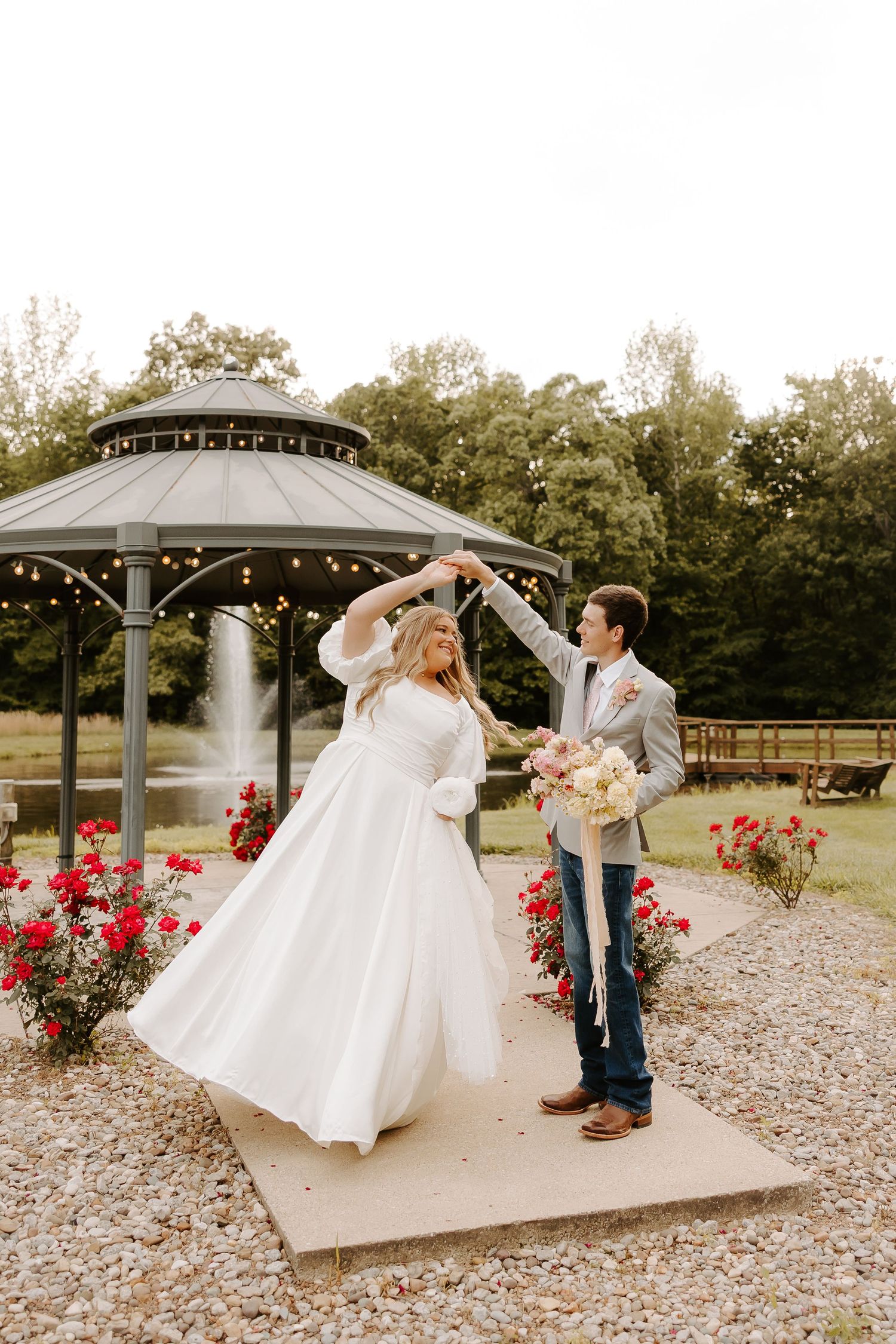 Bride in white gown twirls with groom in gray suit near a gazebo with string lights and red roses.