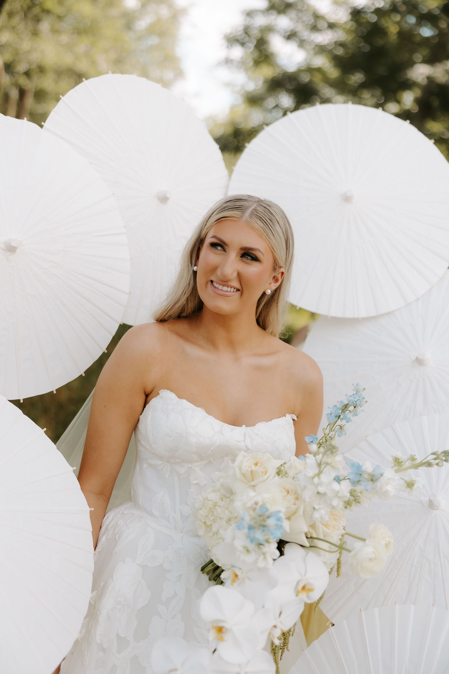Smiling bride in strapless white gown holds floral bouquet surrounded by white paper parasols outdoors.