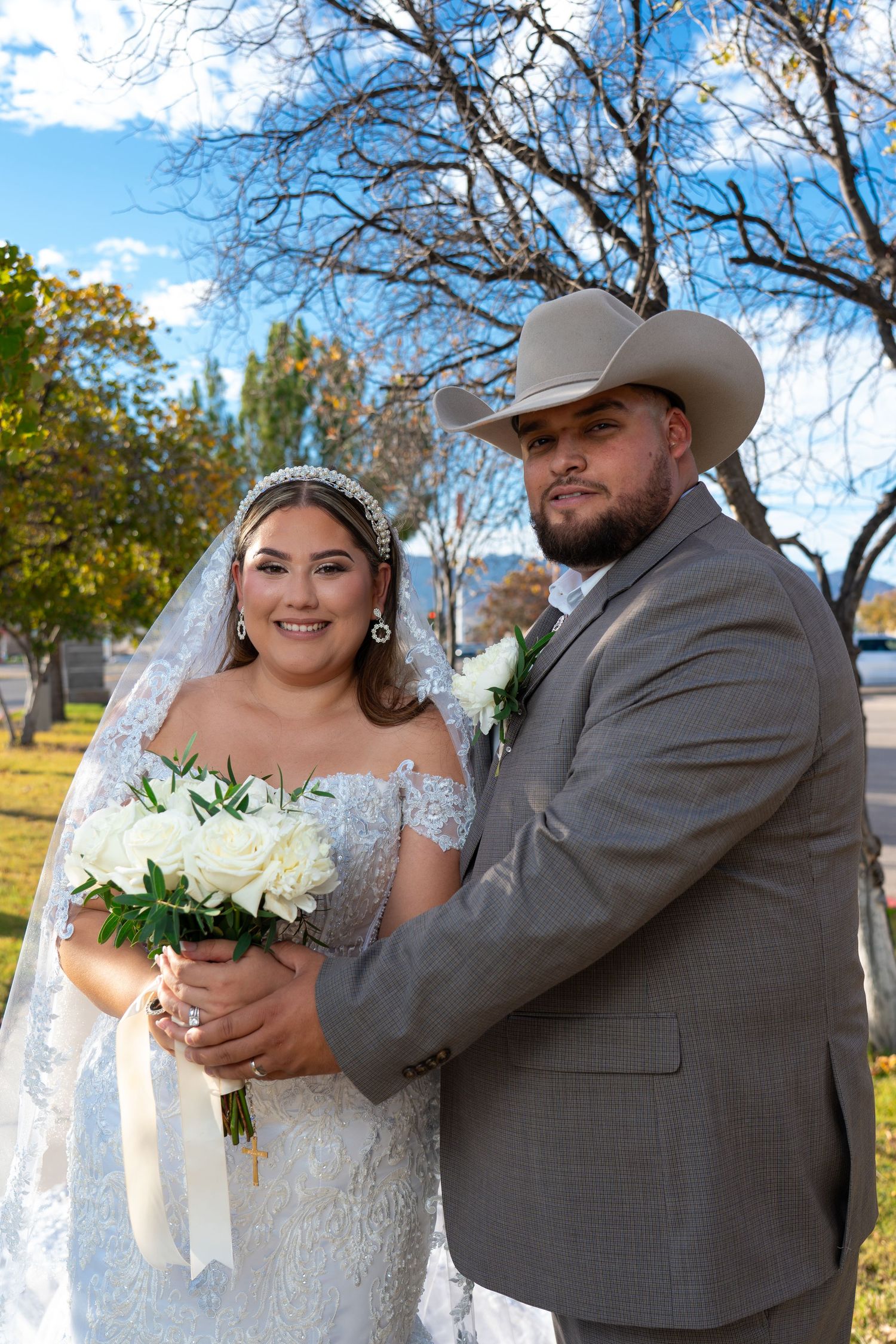 A couple in wedding attire embraces outdoors on their special day, with the bride in white and groom in gray.