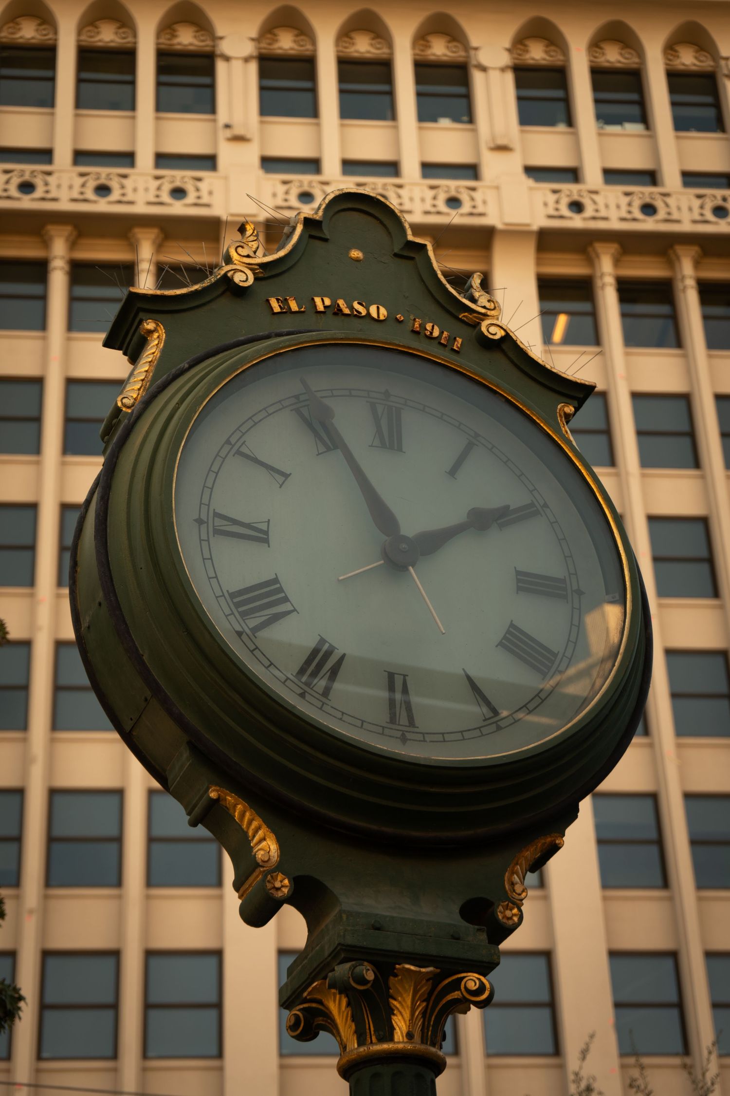 Ornate vintage street clock with Roman numerals mounted on decorative post against backdrop of classical building facade.