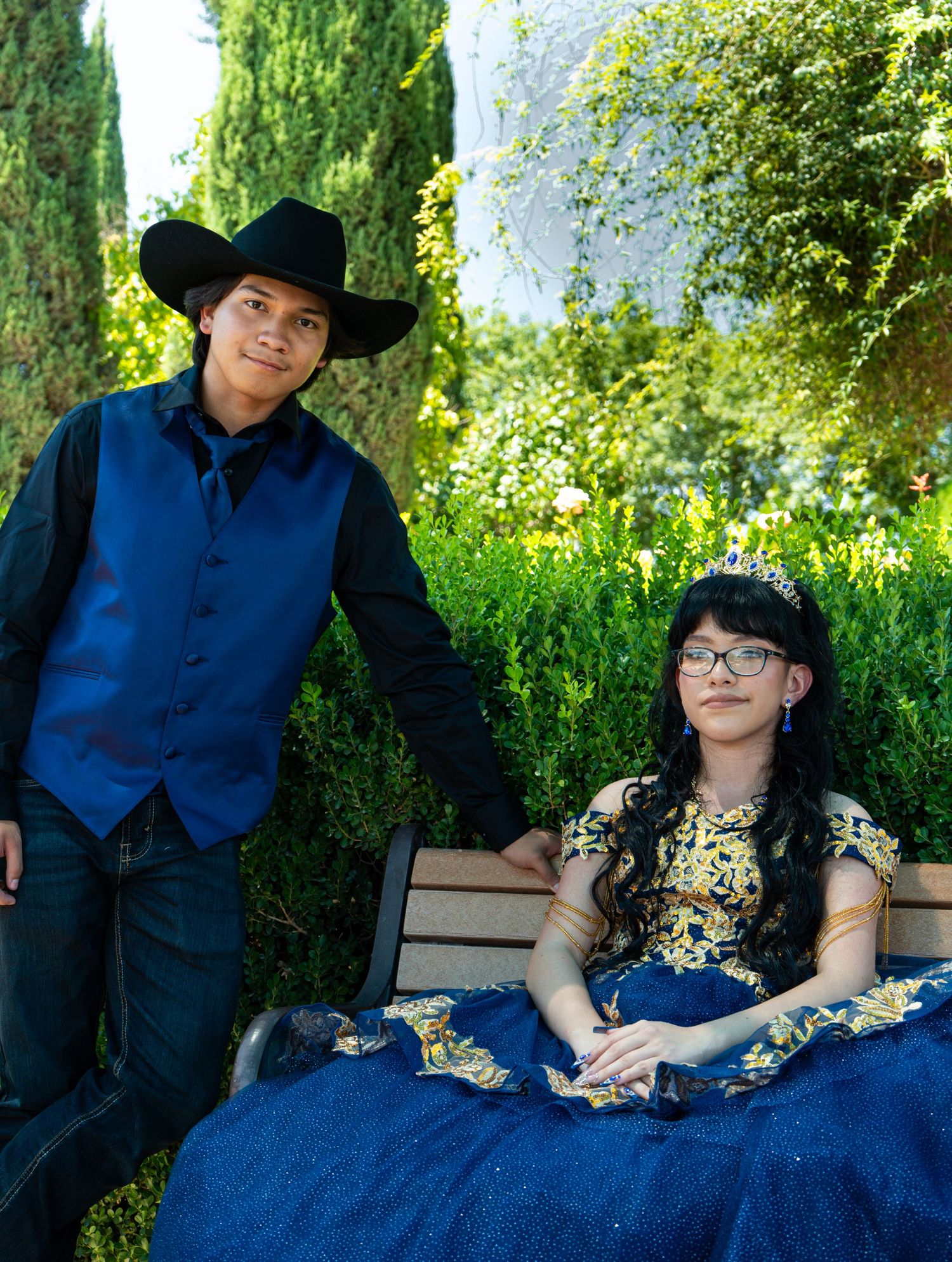 Quinceanera  in western formal wear poses in a lush garden setting with blue and gold accents.