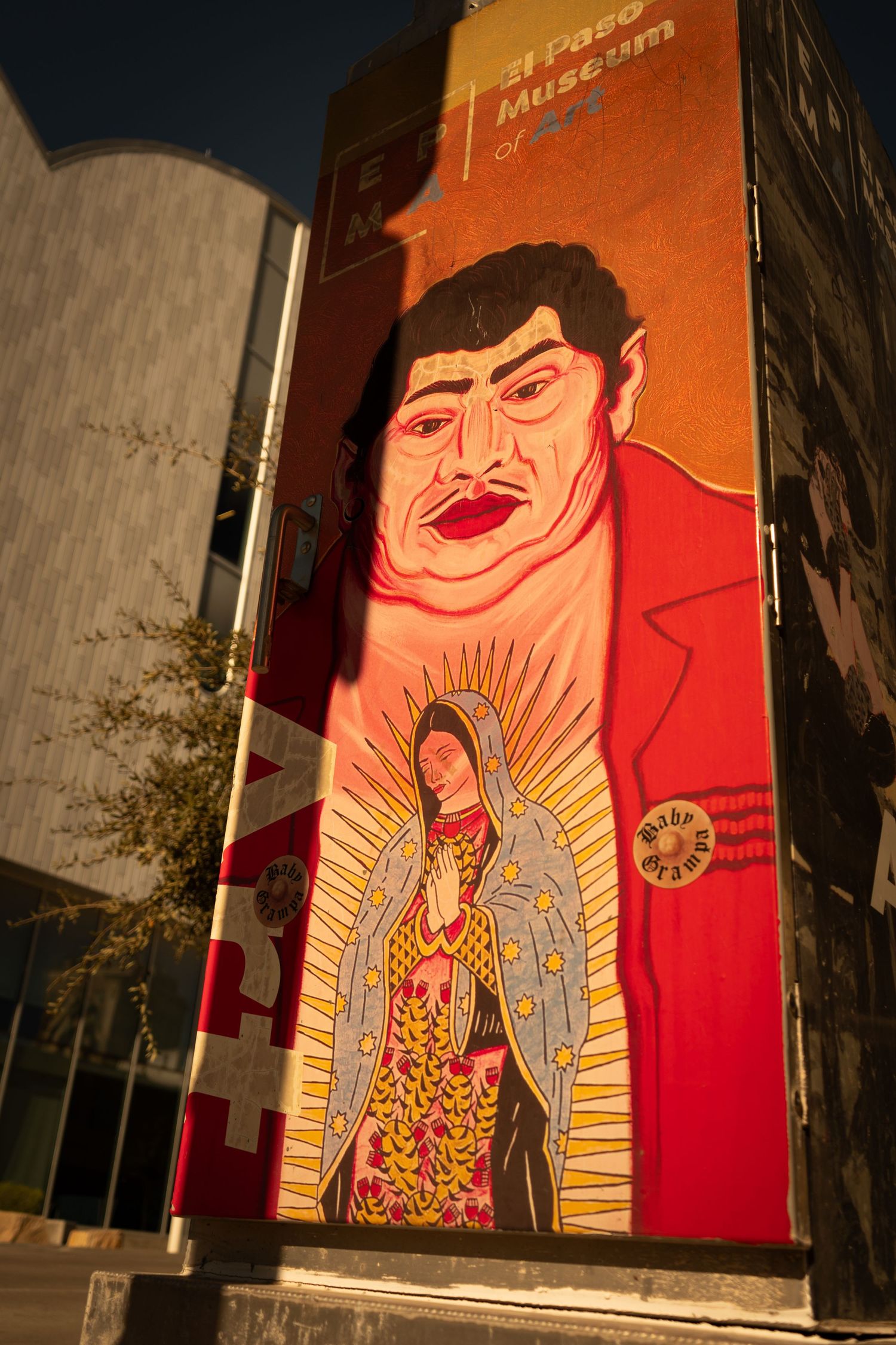 Large street art mural featuring vibrant orange and red colors with a religious figure glowing in golden light against a building wall.