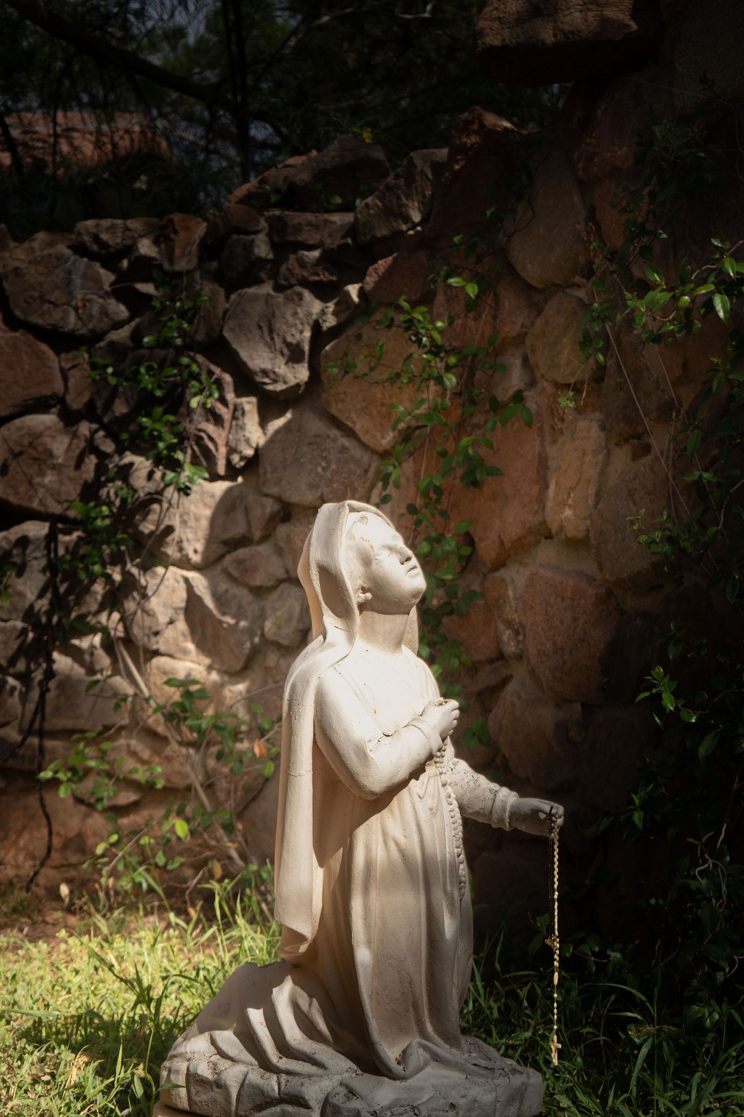 White marble statue in a garden setting illuminated by natural light against a dark stone wall with green foliage.