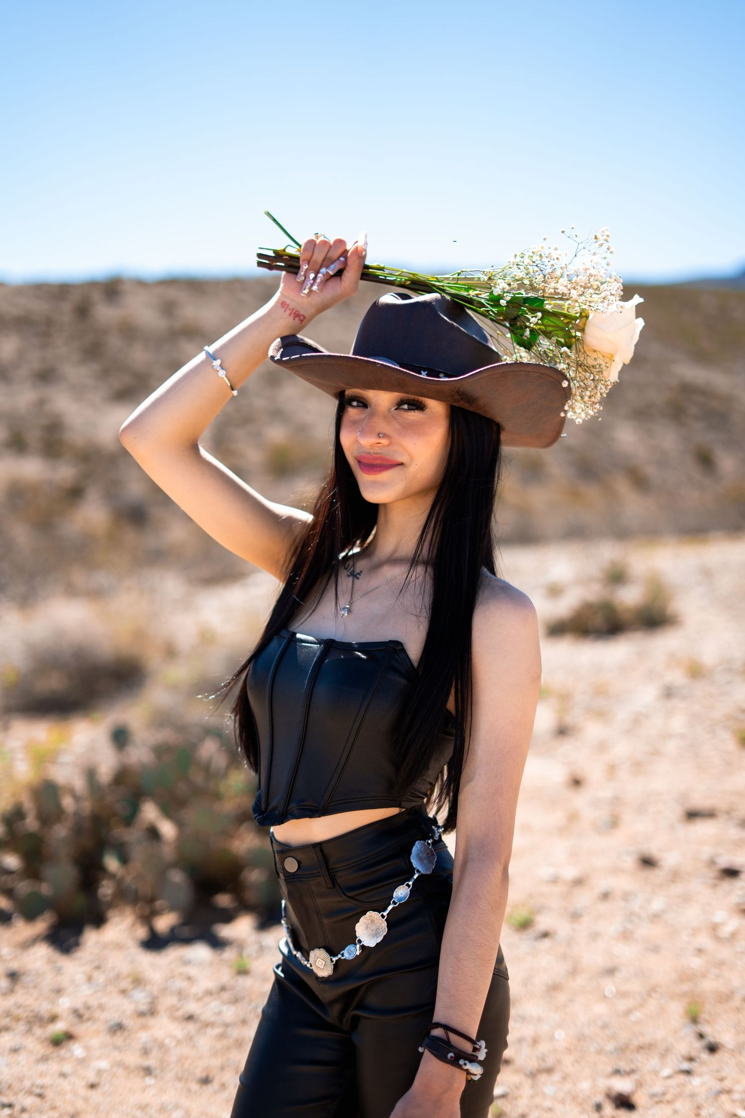 A person in a black dress and wide-brimmed hat poses in a desert landscape against a bright blue sky.