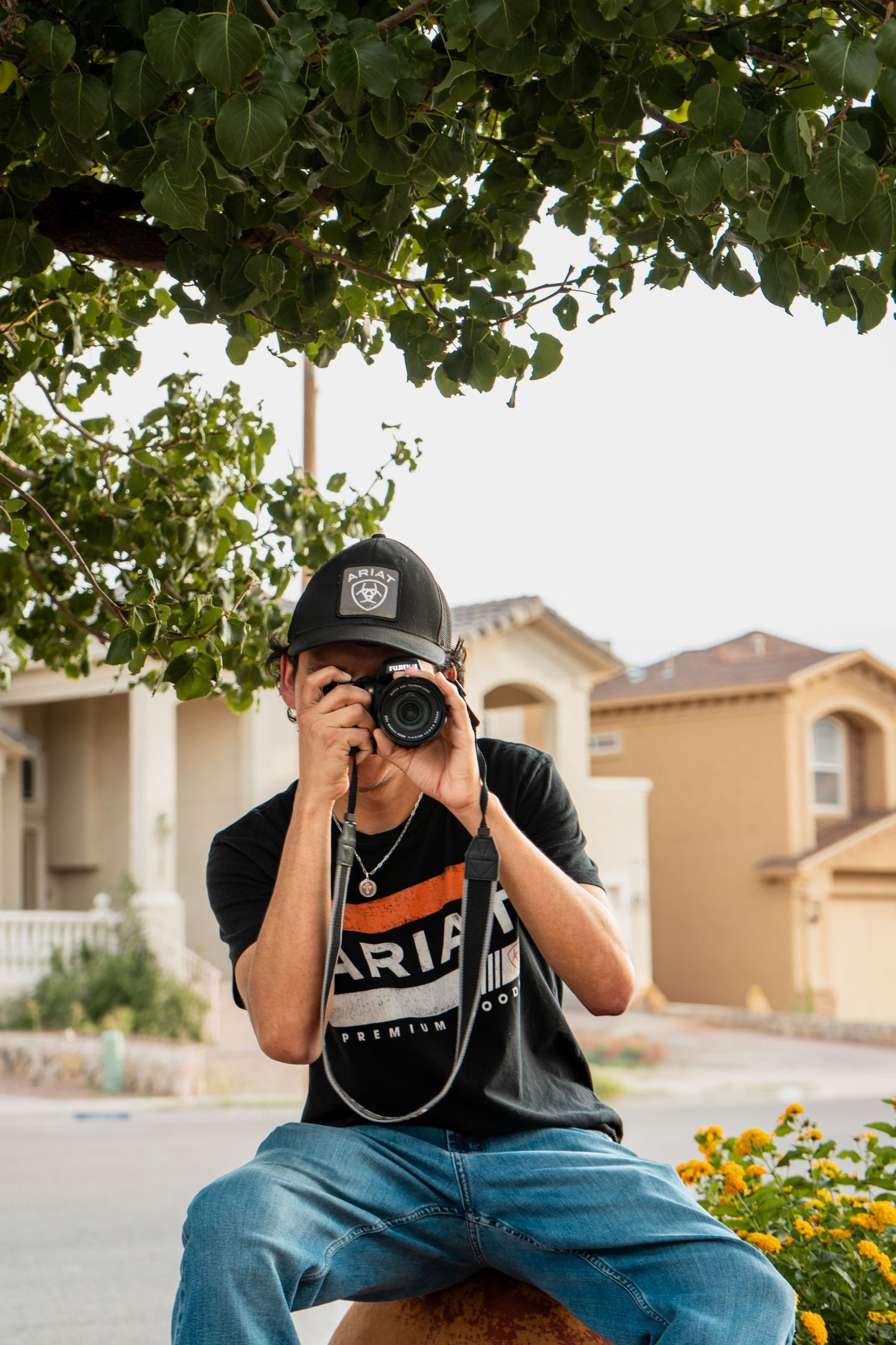 Photographer crouching with camera under green tree foliage in suburban setting.
