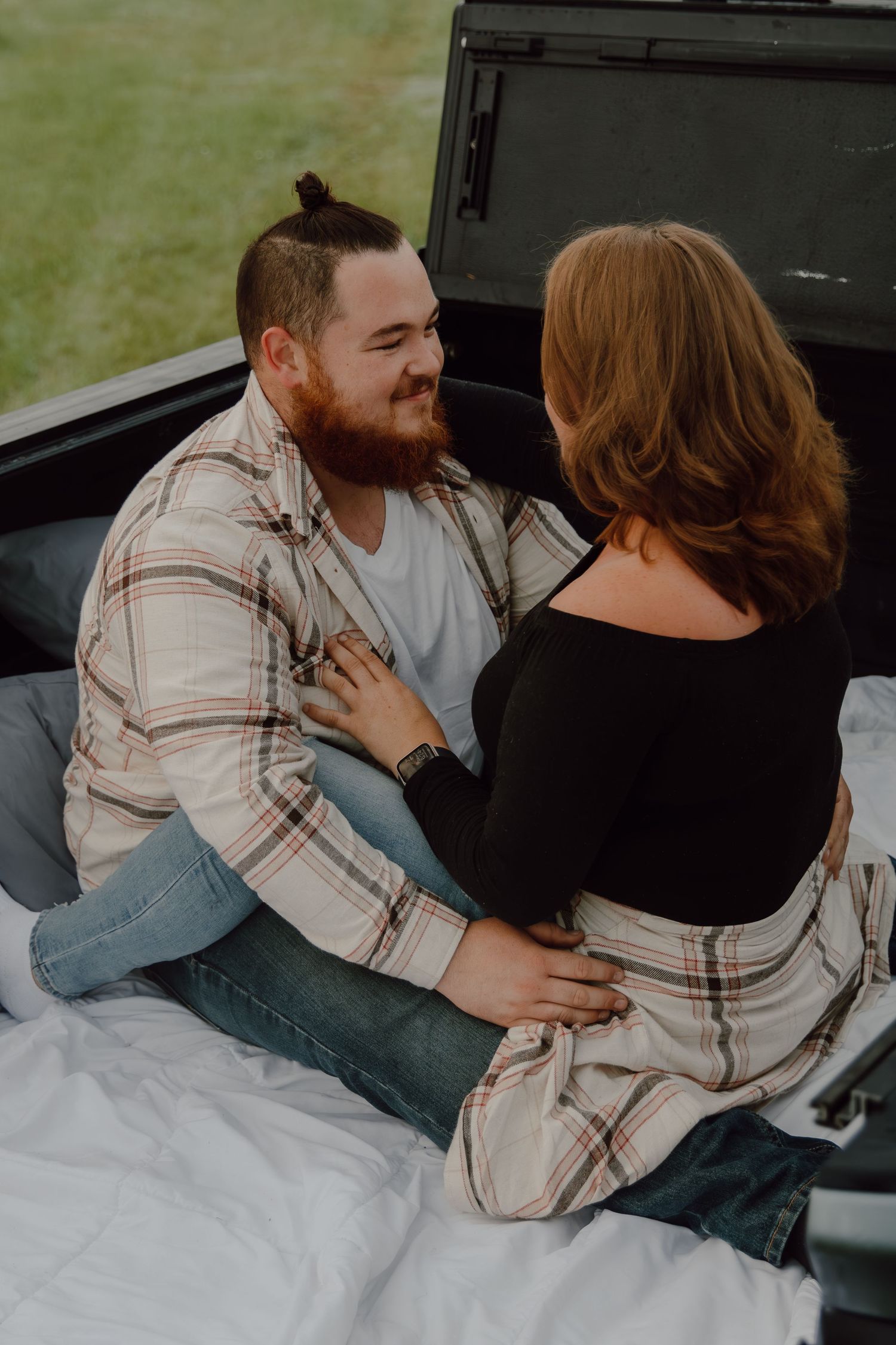 A bearded man in a plaid shirt smiles at a redhead woman sitting together in a truck bed outdoors.