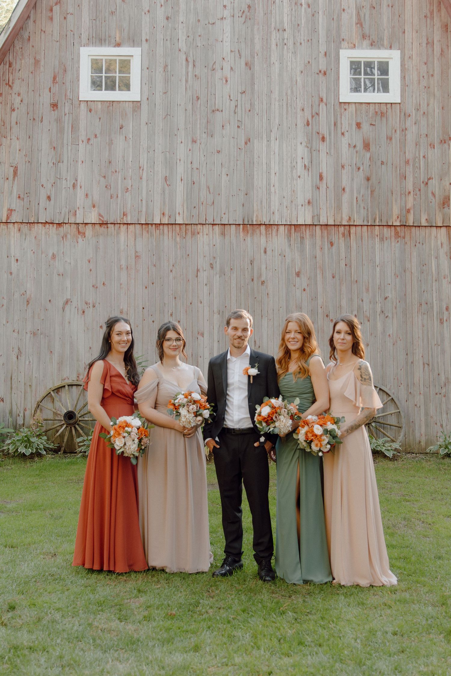 Wedding party poses in mix-matched dresses in earth tones against a rustic barn backdrop.