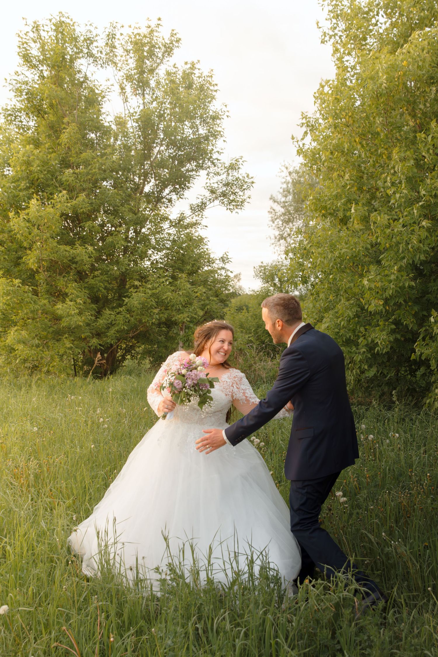 A romantic wedding couple dances playfully in a field with tall grass and trees at golden hour.