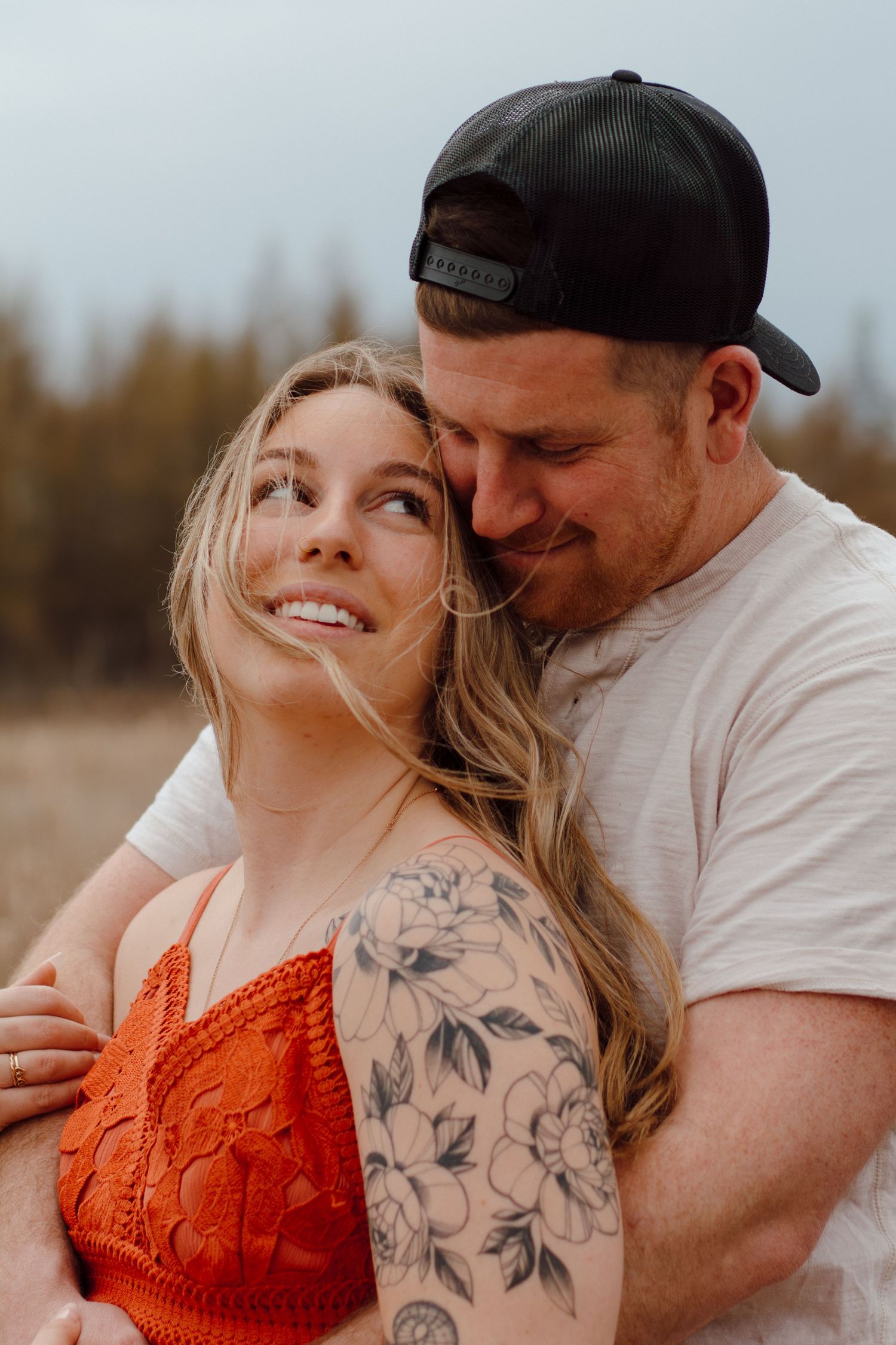 Couple embracing outdoors, woman in red lace top with floral arm tattoo smiling up at man in backward cap.