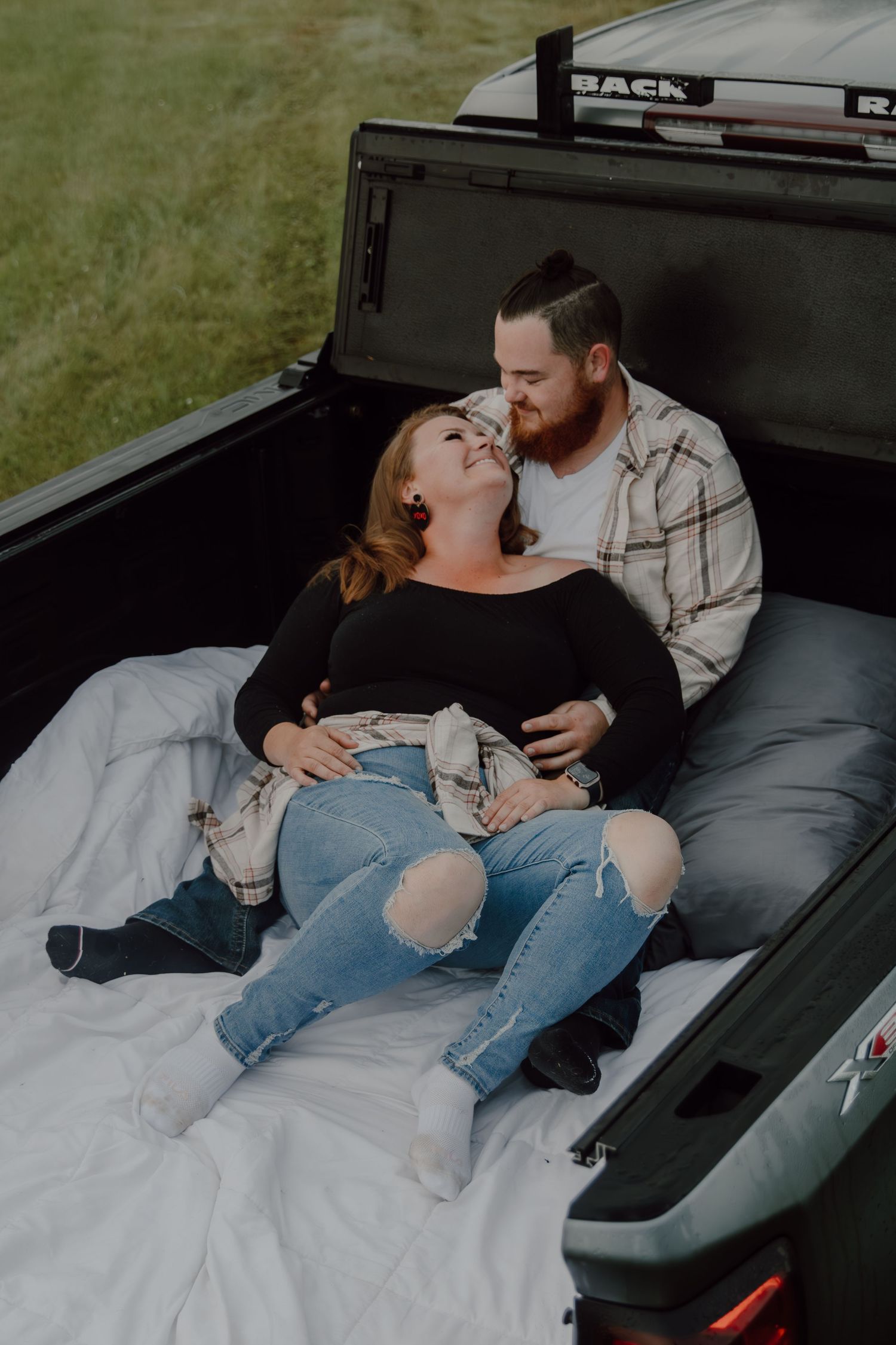 Couple relaxing together in the bed of a pickup truck, sharing a tender moment on a white blanket outdoors.